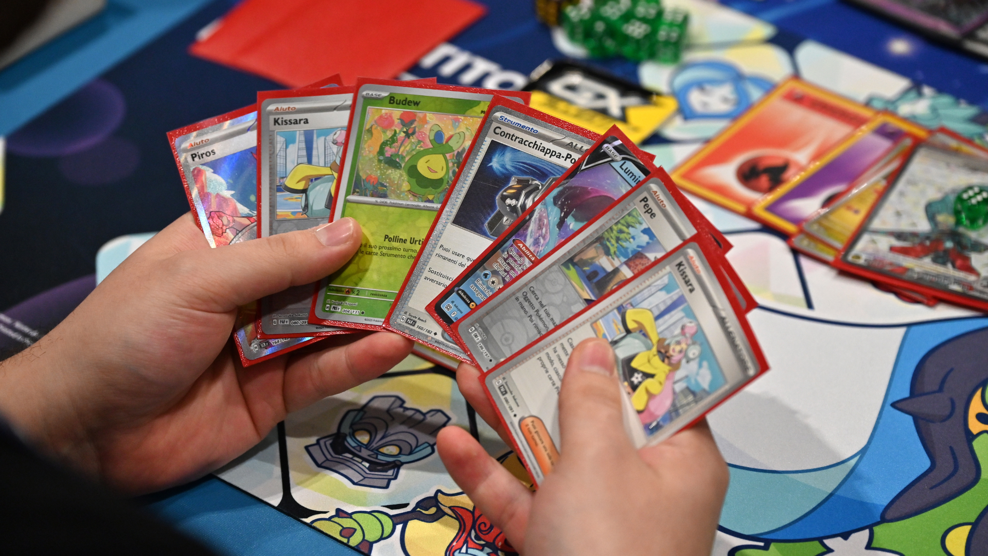 A person holding a hand of Pokémon cards during a game, with a colorful playmat and dice visible in the background.