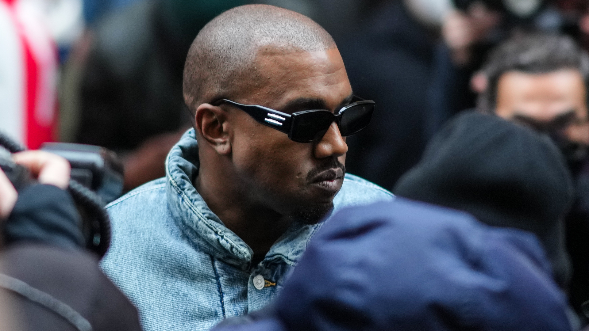 Kanye West wearing sunglasses and a denim jacket, surrounded by people and photographers.