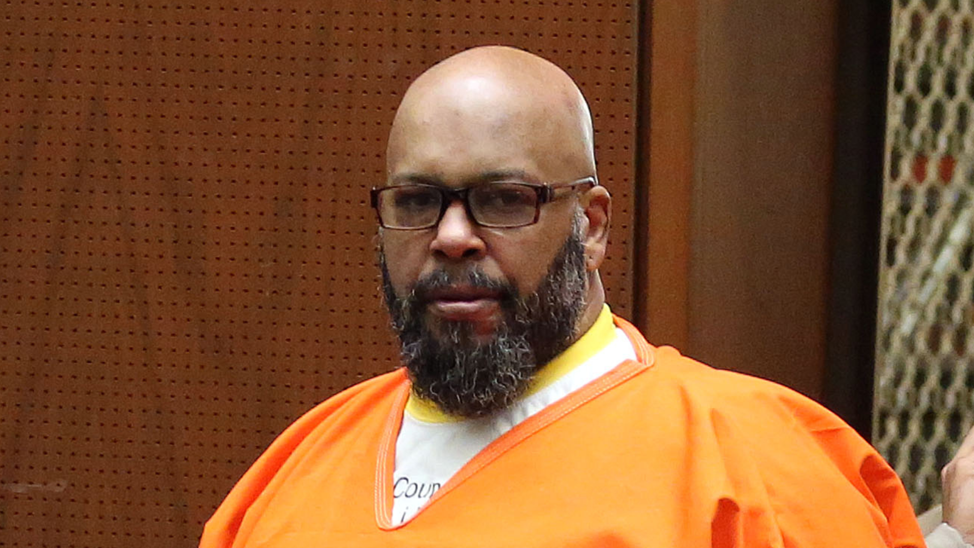Suge Knight with a beard and glasses wearing an orange prison jumpsuit in a courtroom.
