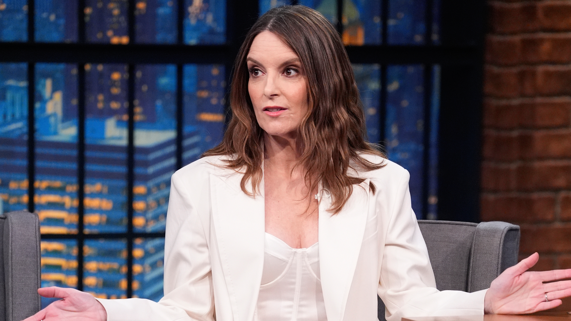 Tina Fey in a white outfit, gesturing with her hands, sitting on a talk show set with a cityscape background.