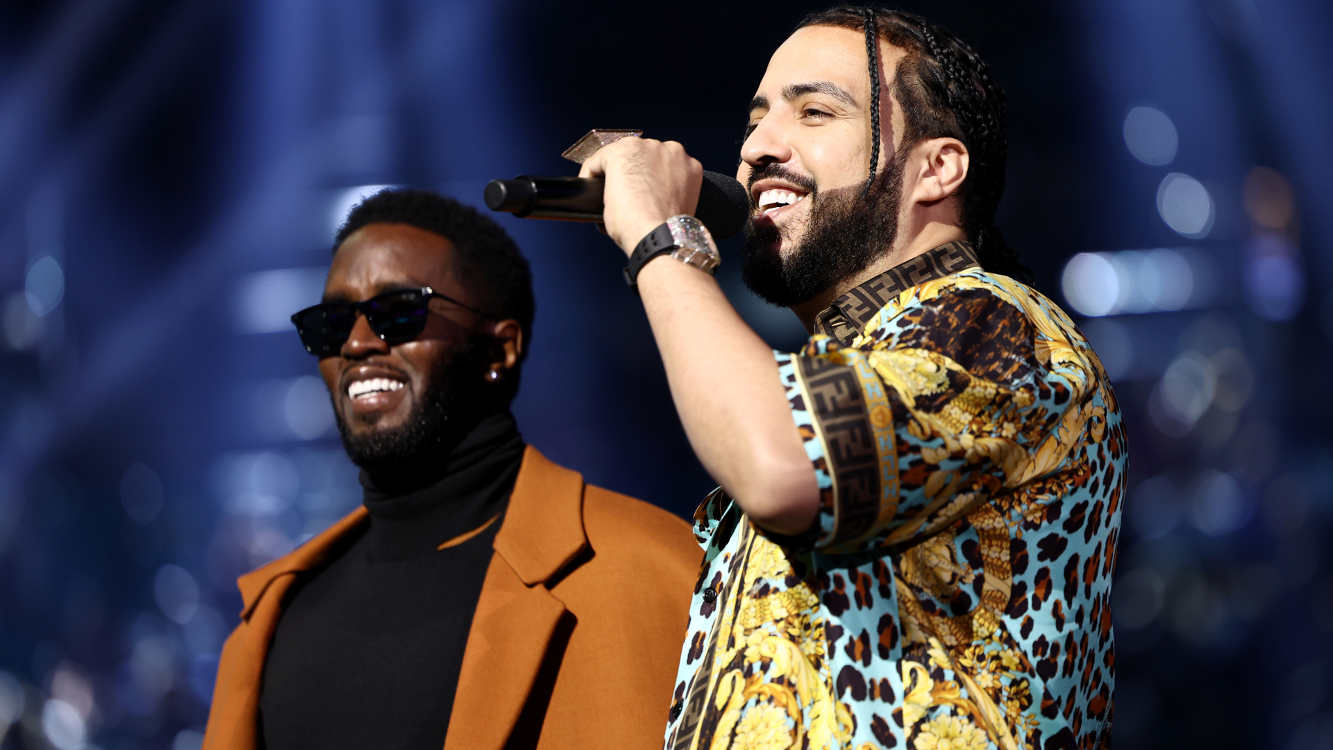 Diddy in sunglasses and a brown coat stands beside French Montana, who is holding a microphone and wearing a patterned shirt.