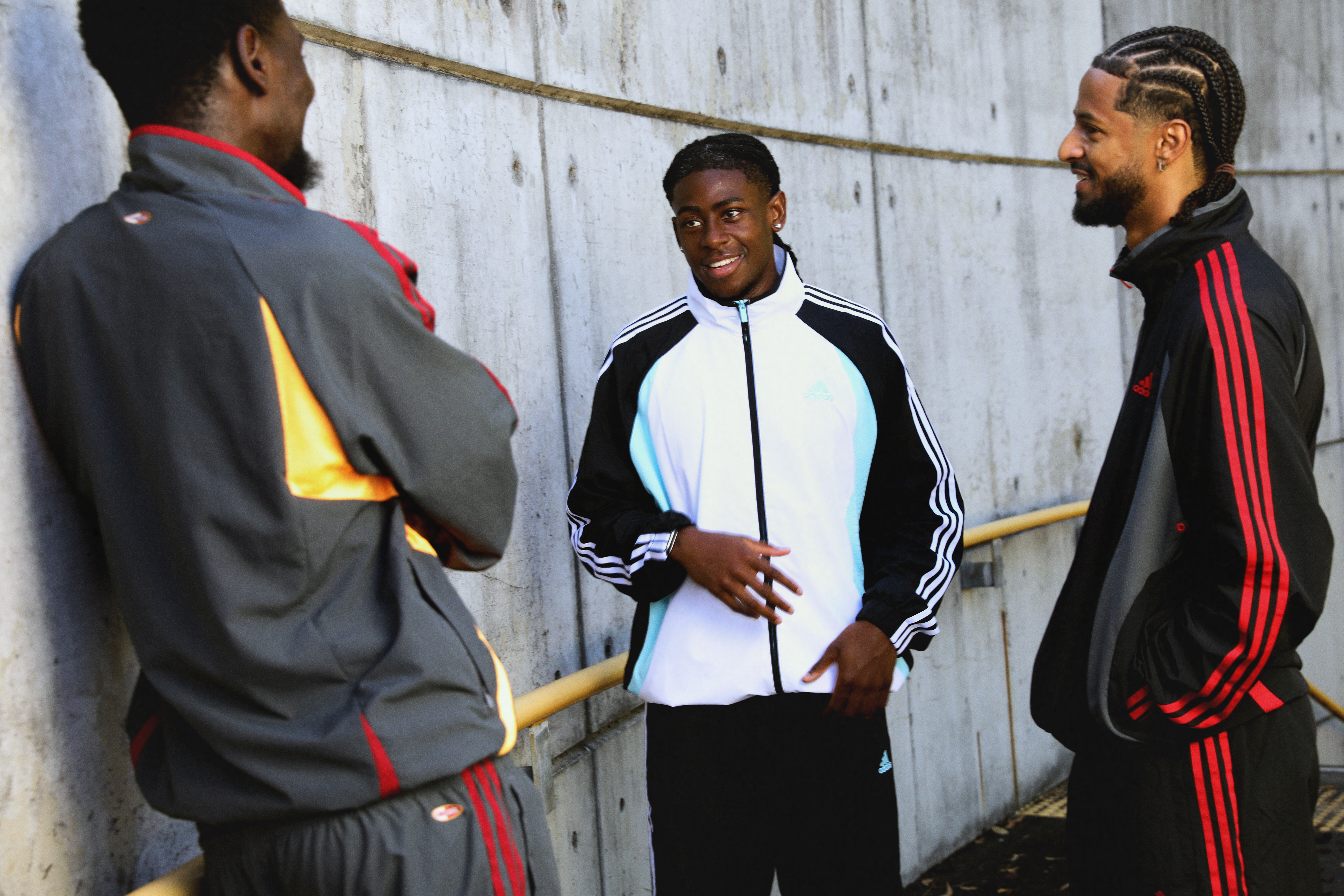 Three men in sportswear converse outdoors, standing by a concrete wall. They appear relaxed and engaged in conversation.