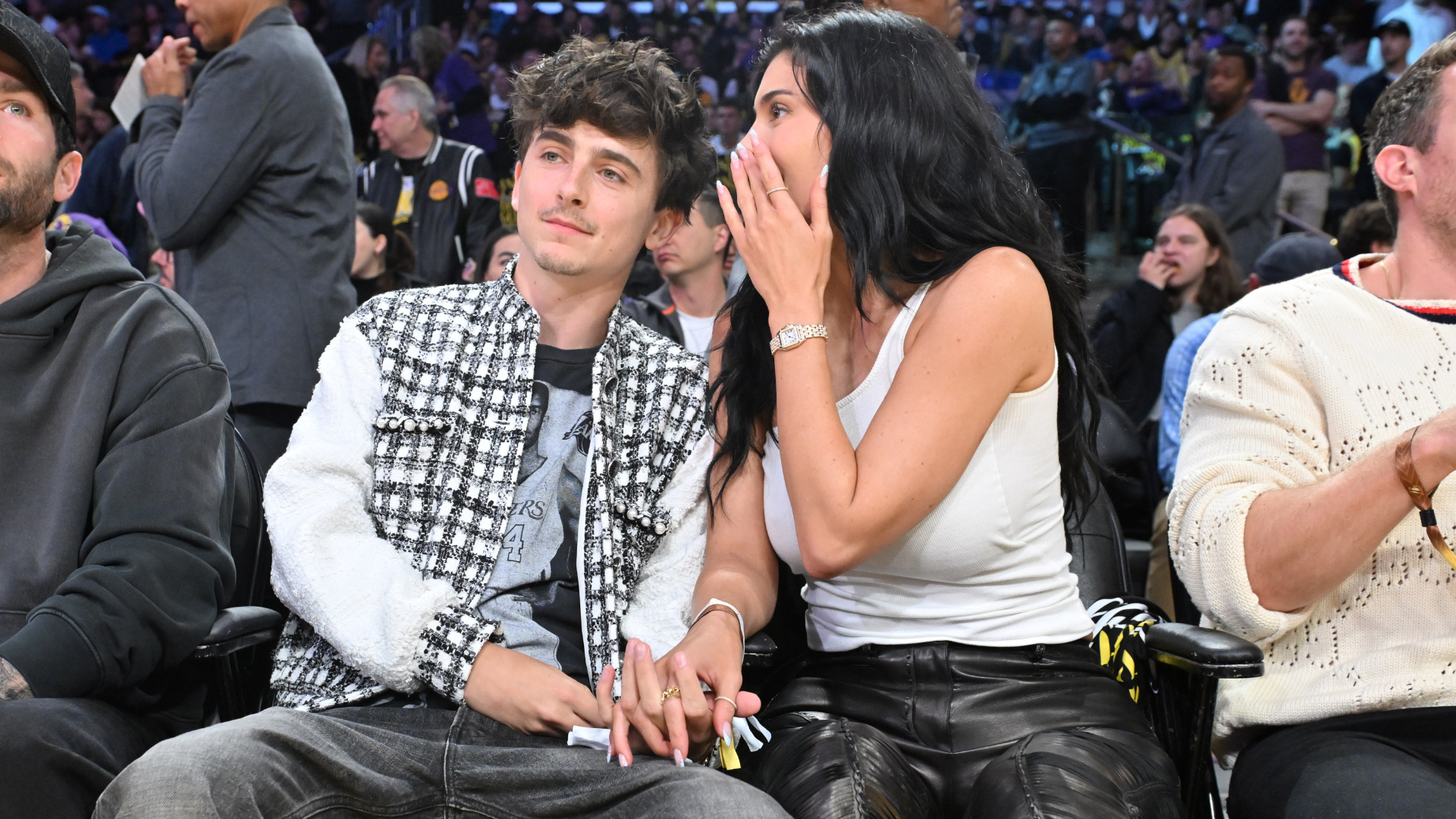 Timothée Chalamet and Kylie Jenner sit courtside at a basketball game, holding hands. Kylie whispers to Timothée.