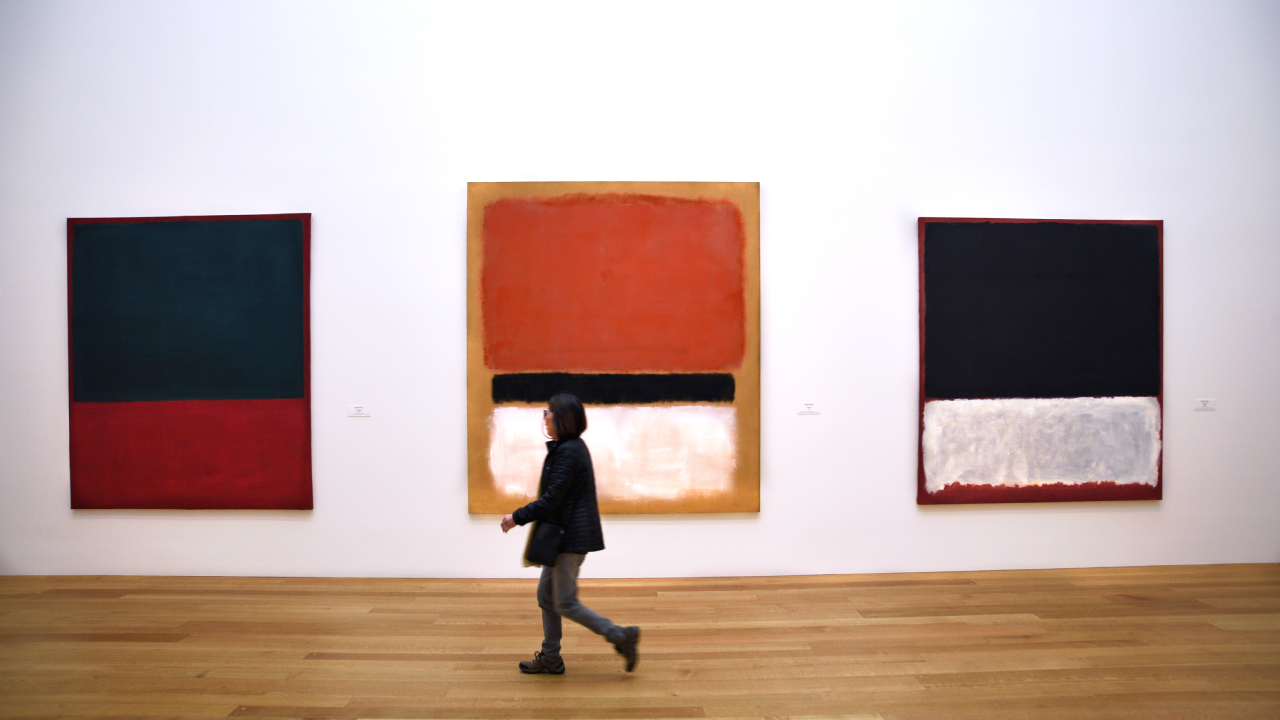 A person walking past three abstract paintings by Mark Rothko in a gallery. The artworks feature large blocks of color.