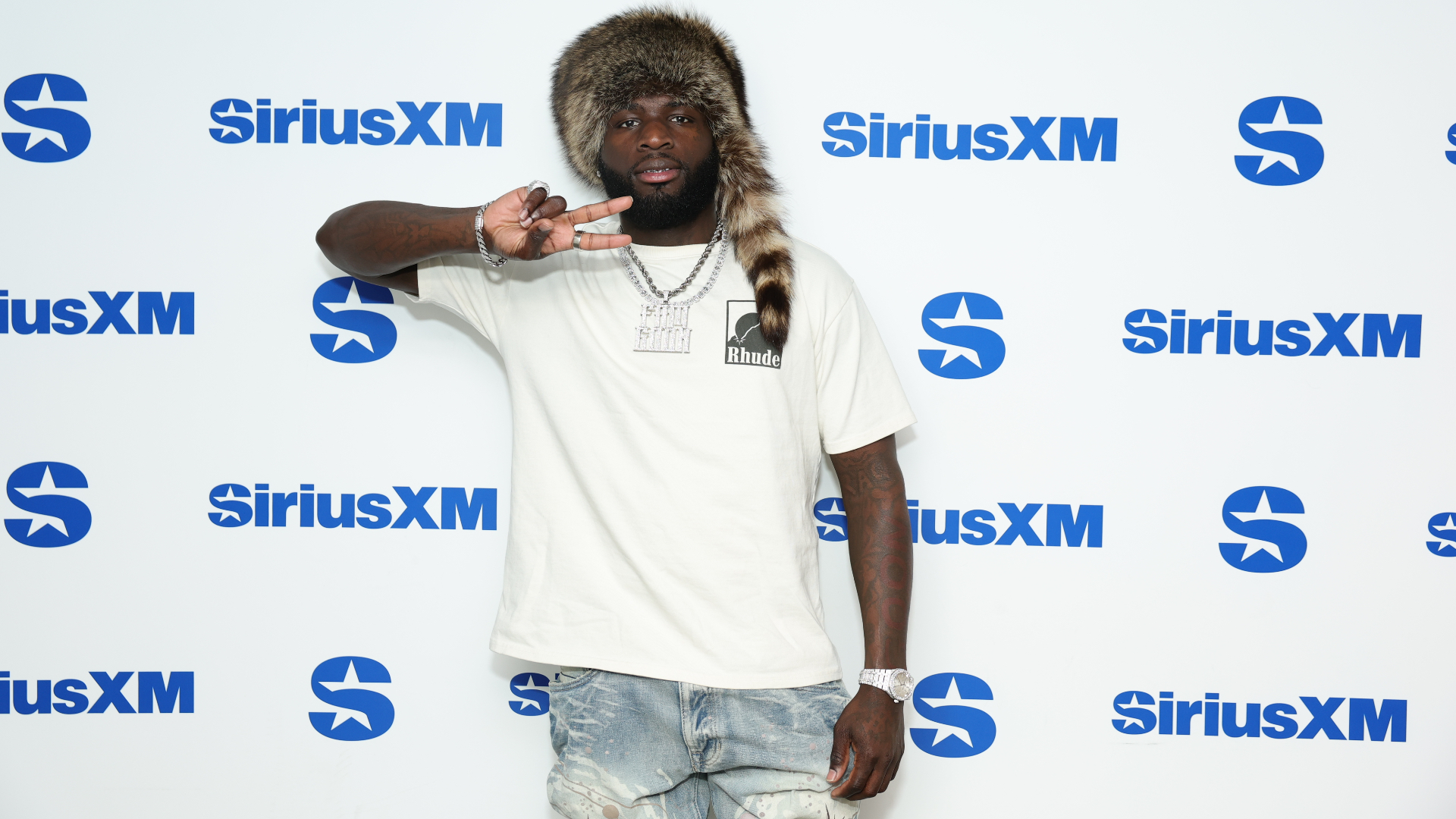 A man wearing a fur hat and white t-shirt poses in front of a SiriusXM backdrop, making a peace sign with his fingers.