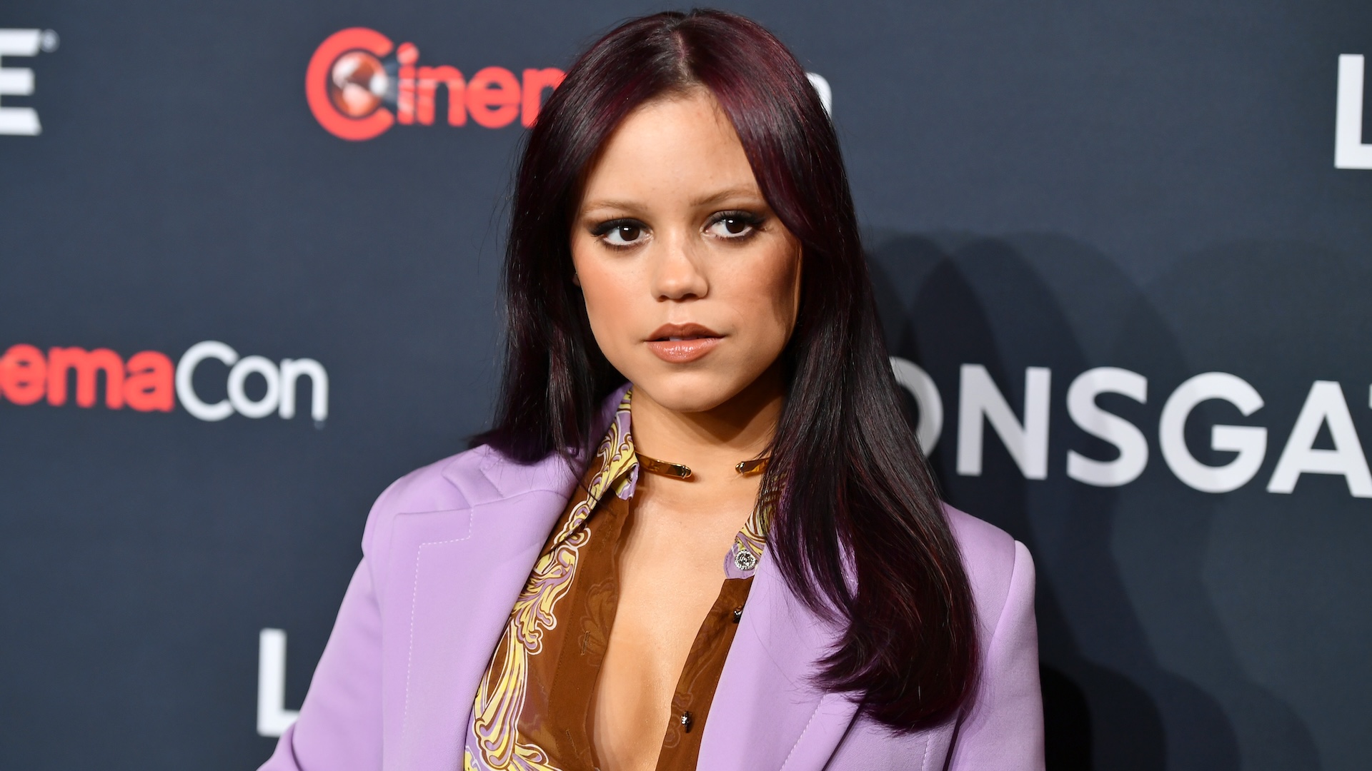 Jenna Ortega at a CinemaCon event, wearing a purple blazer and patterned shirt, standing against a black backdrop with logos.