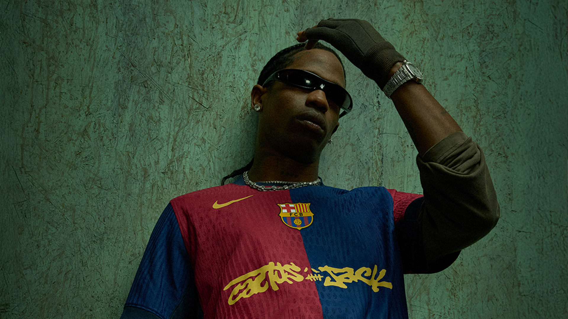 Travis Scott wearing sunglasses and a Barcelona jersey, posing with one hand on their head against a textured background.