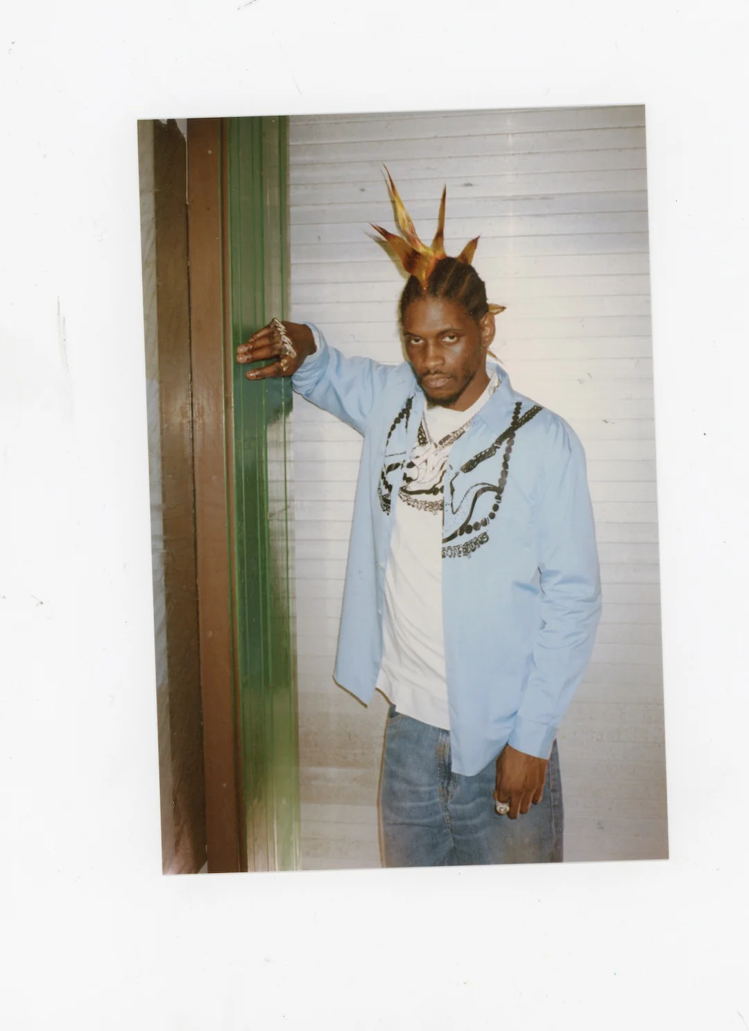 ASAP Nast with unique spiked hairstyle, wearing a light blue shirt over a white t-shirt and jeans, leaning against a wall.