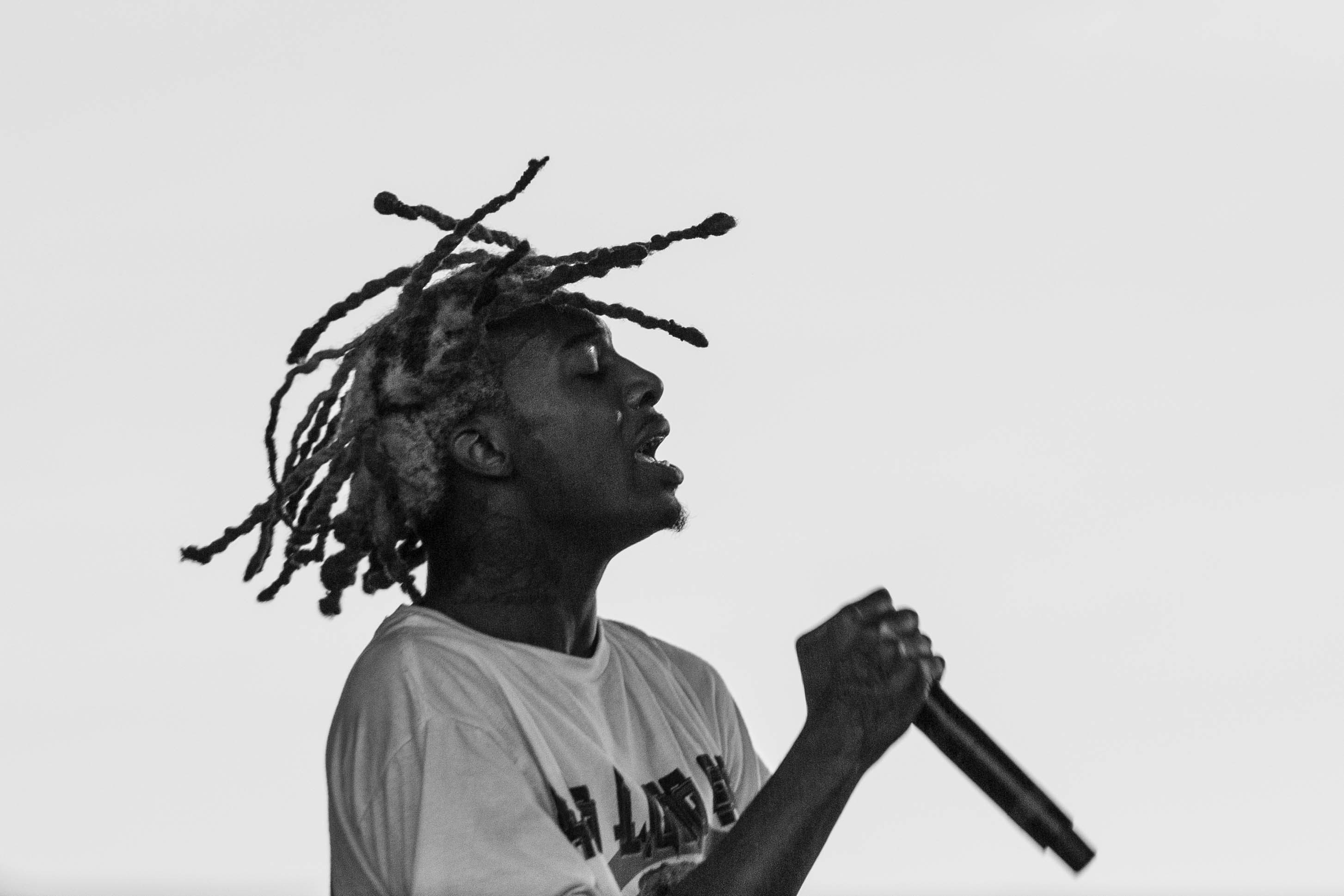 A black and white image of Playboi Carti with distinctive dreadlocks performing on stage, holding a microphone and wearing a casual shirt.