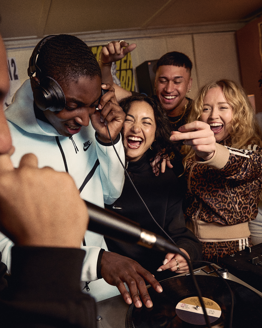 A group of people joyfully gathered around a DJ, who is wearing headphones and spinning vinyl records. They are laughing and pointing.