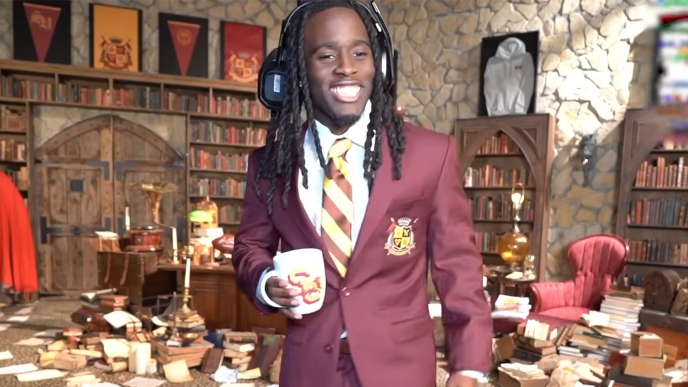 Kai Cenat wearing a maroon blazer and striped tie, holding a mug, stands in a room filled with books and medieval decor.