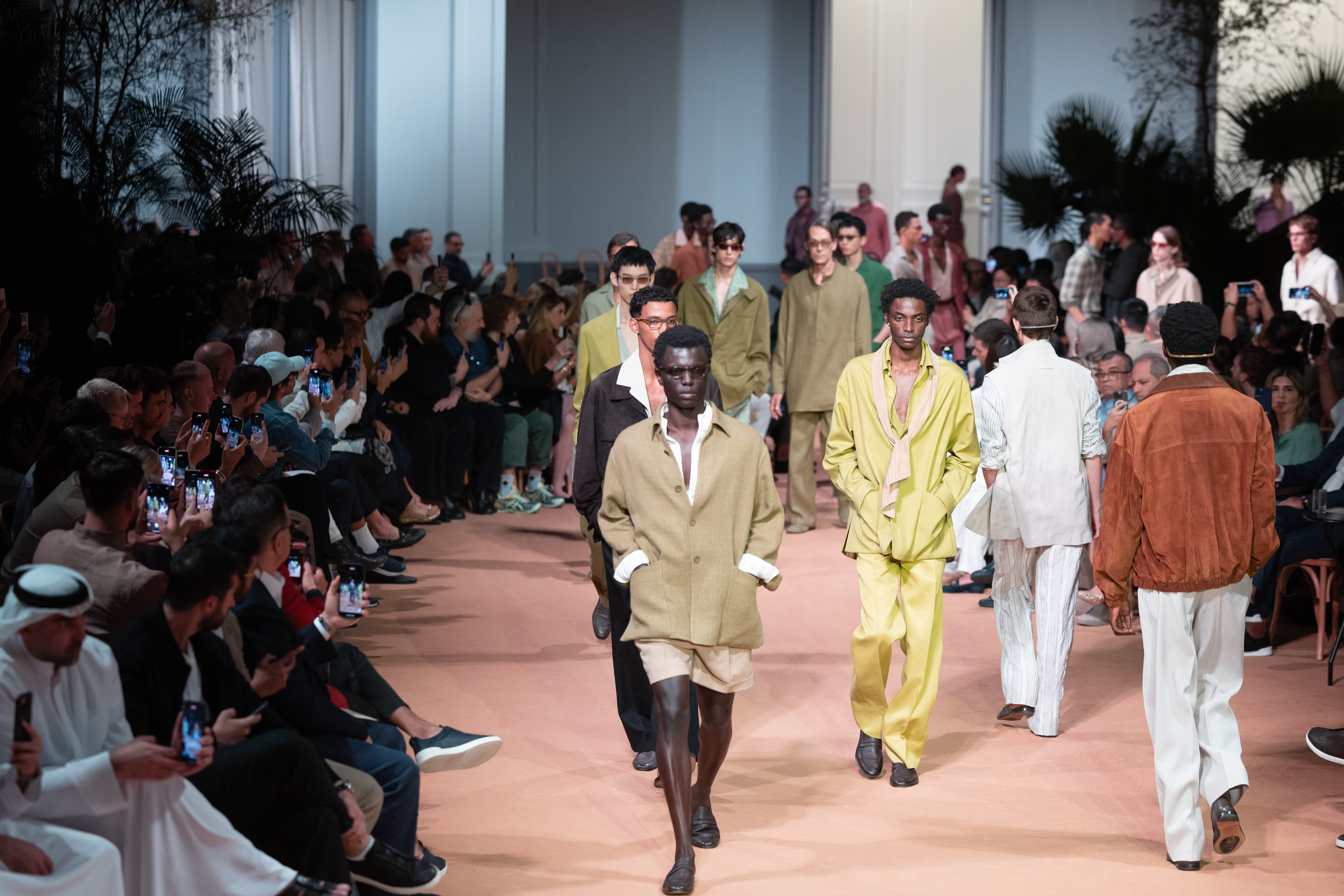 Models walking on a fashion runway, wearing earthy-toned outfits. The audience is seated on either side, capturing the show on their phones.