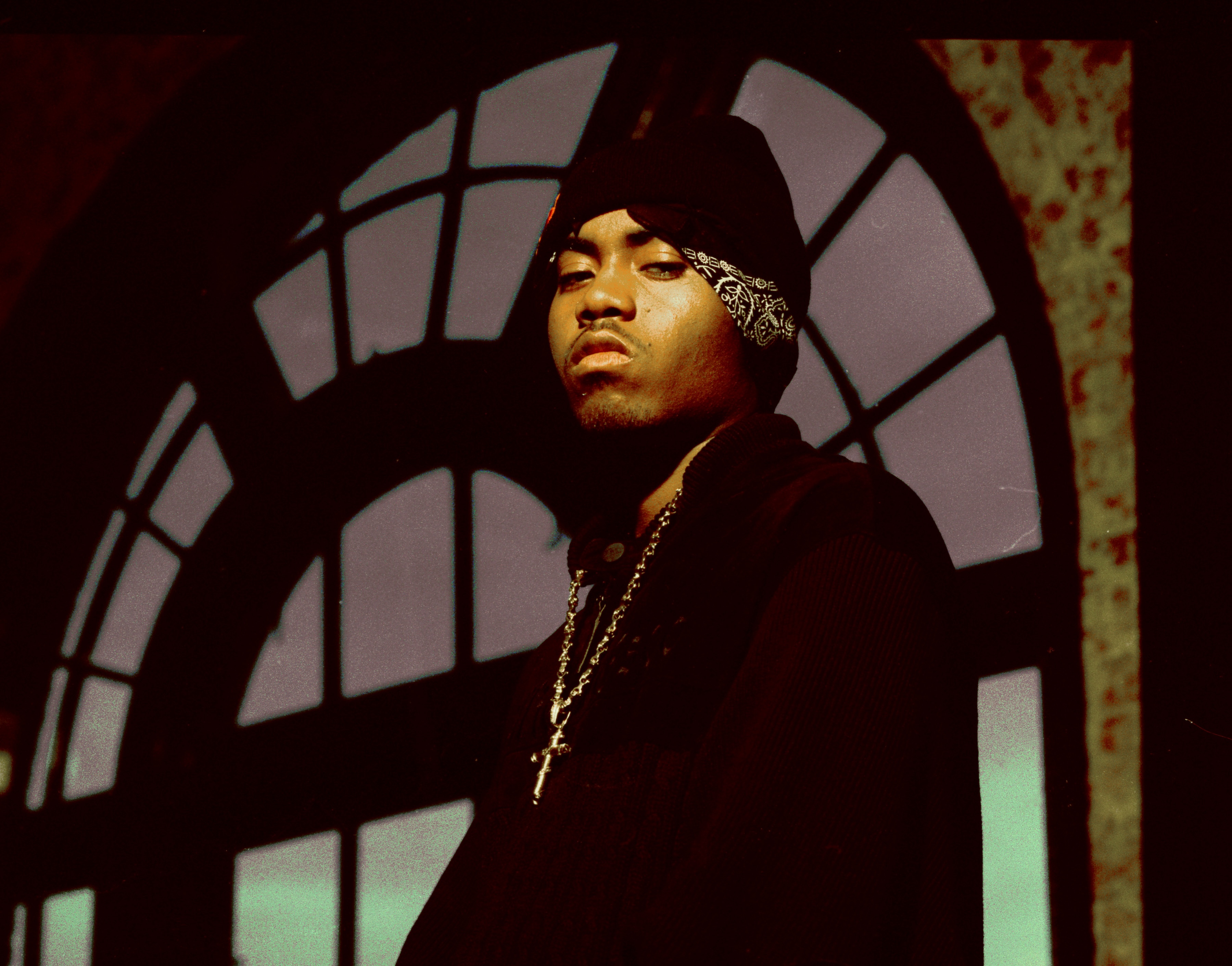 Nas wearing a dark beanie and bandana stands in front of an arched window, looking serious.
