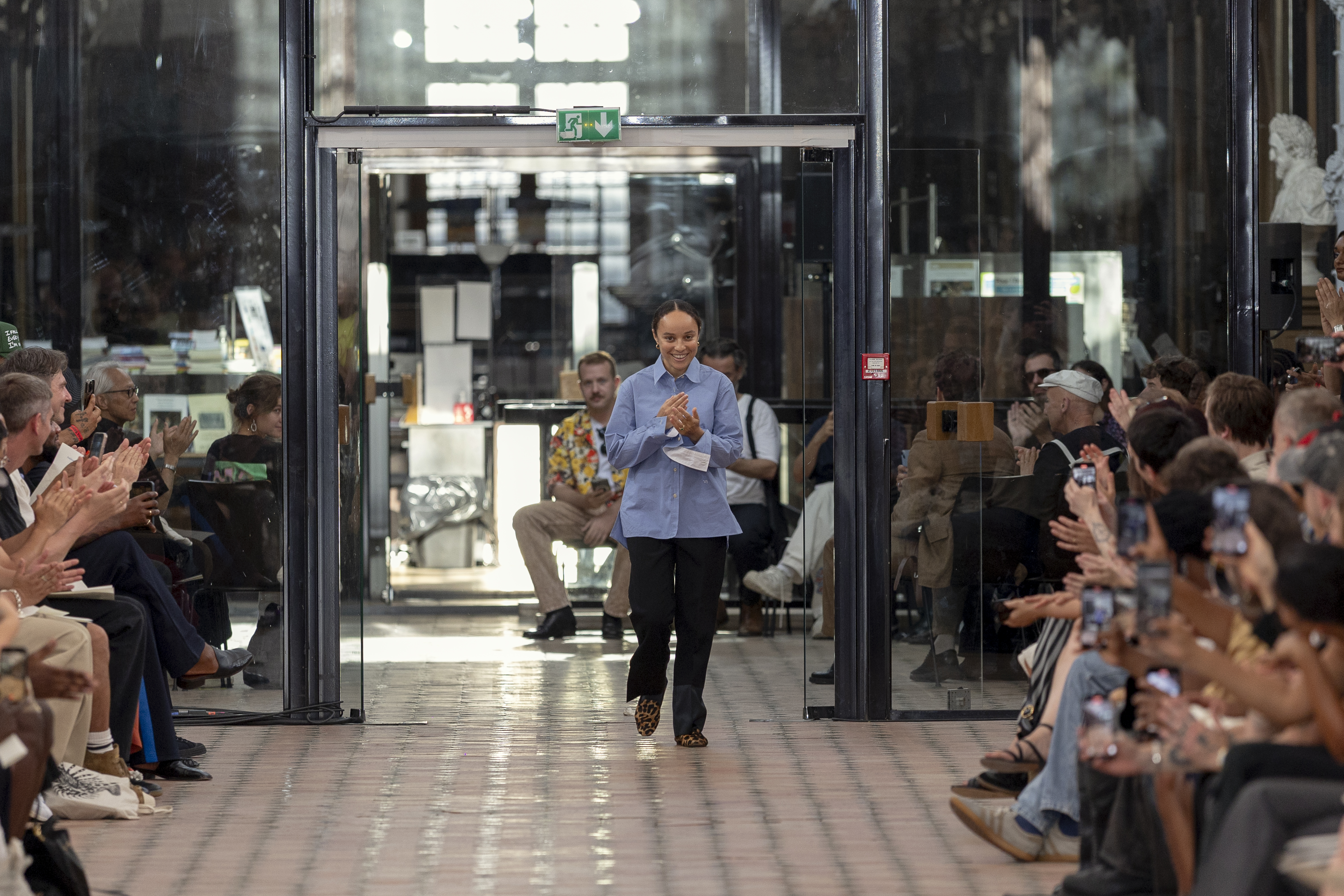 A fashion show runway with a designer walking down the aisle, applauded by the audience. The setting is modern and stylish.