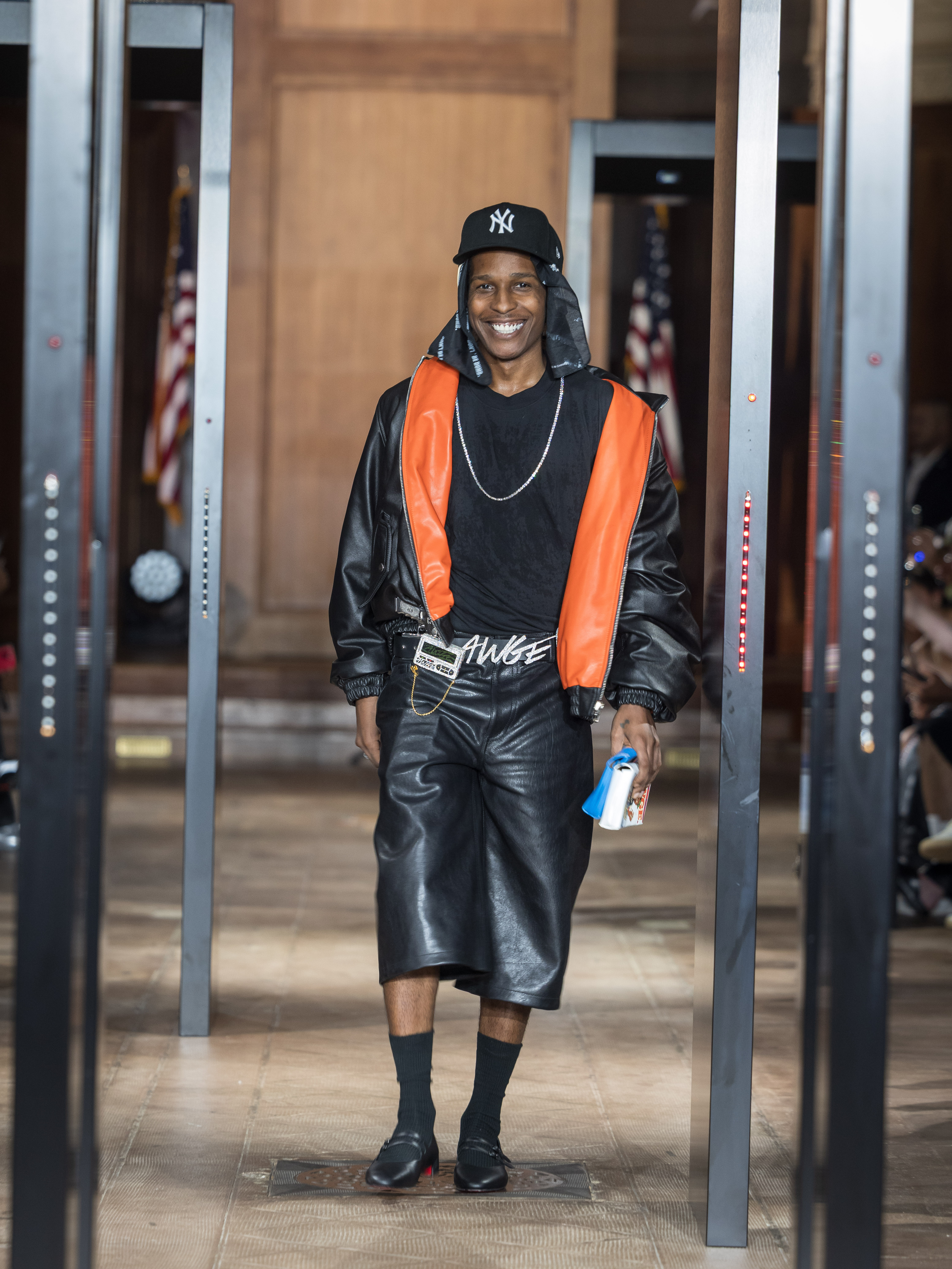 A$AP Rocky walking confidently, wearing a black and orange leather outfit with a New York Yankees cap, smiling.