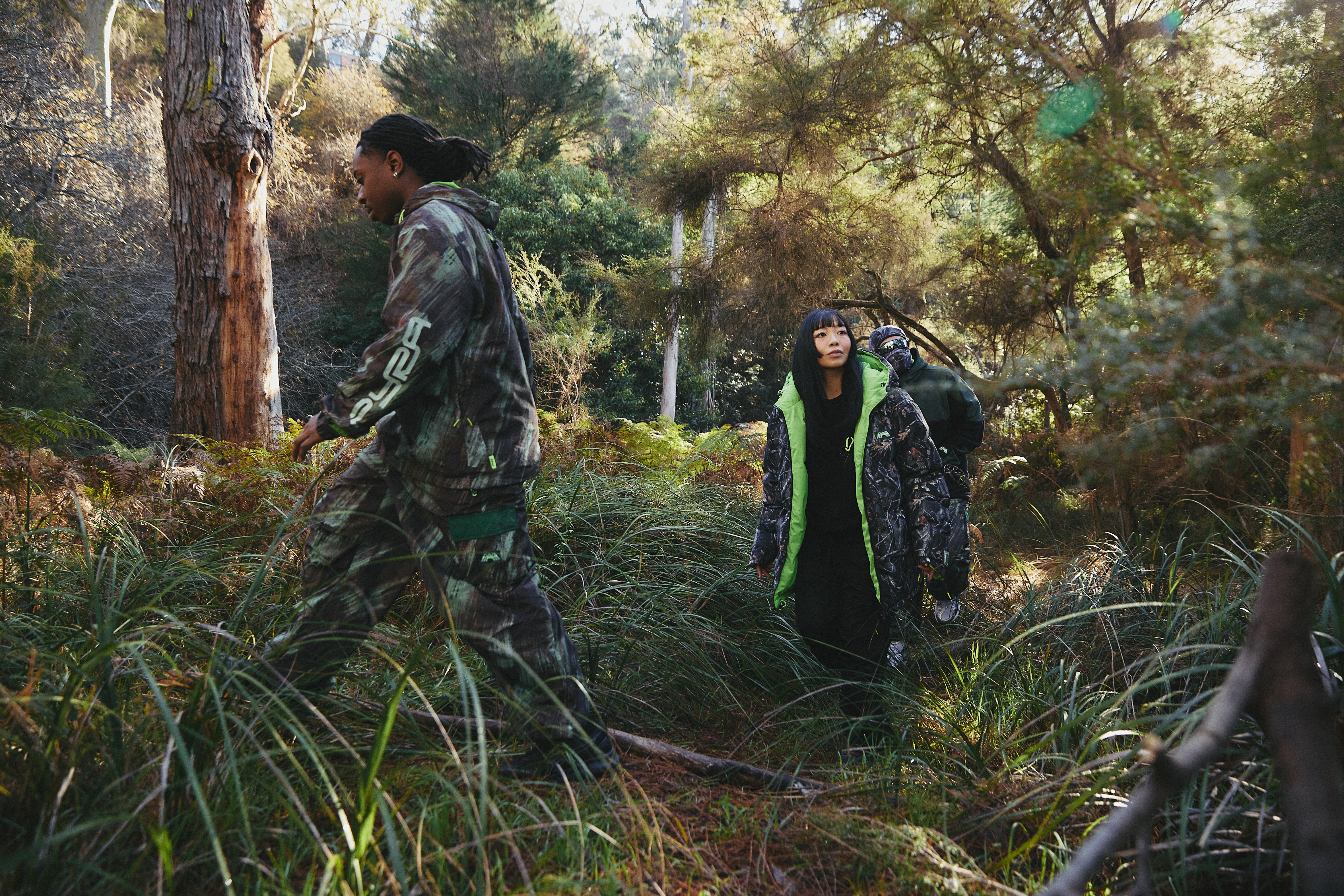 Two people in camouflage clothing walking through a dense forest, surrounded by tall trees and lush greenery.