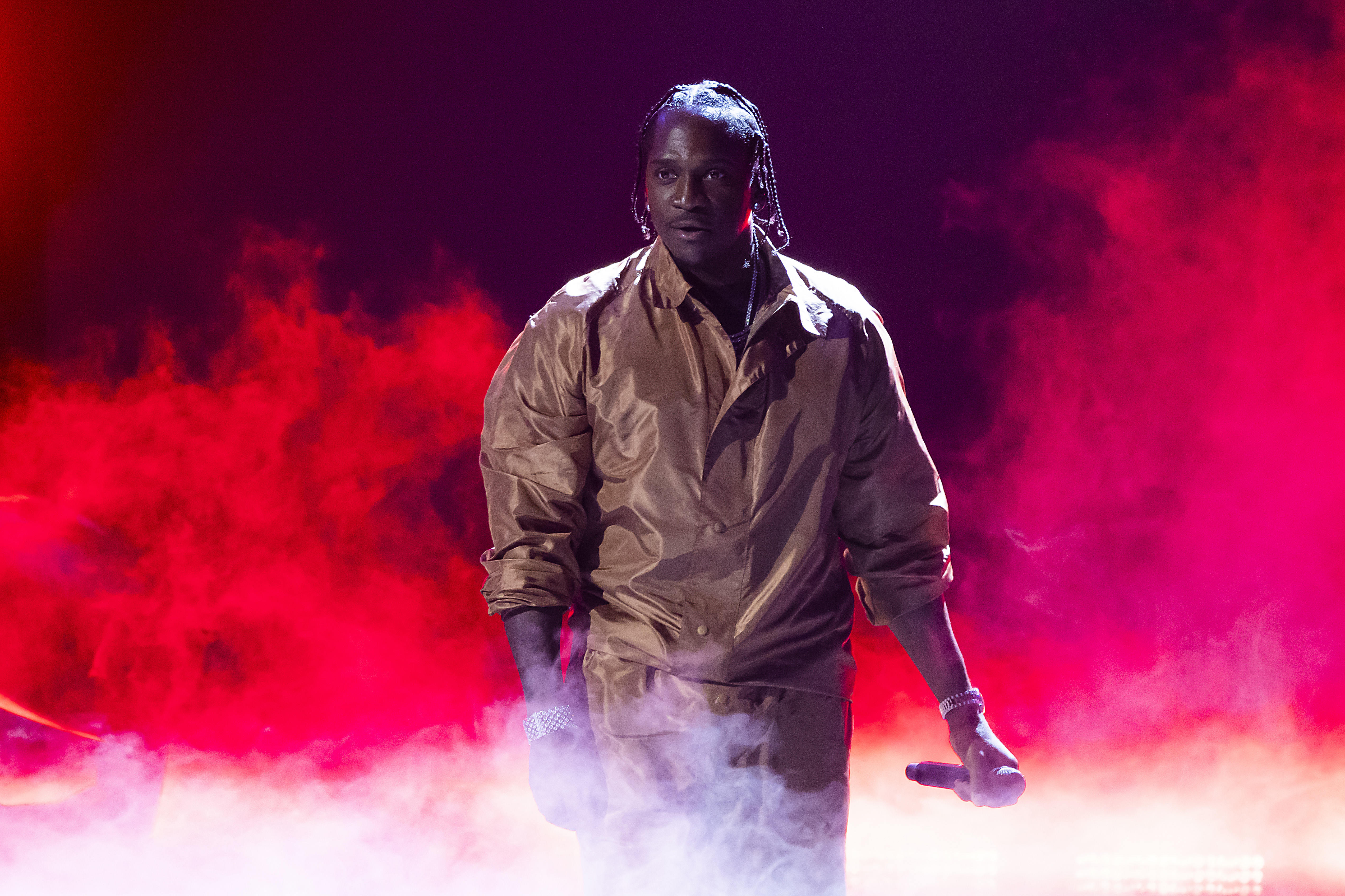 Pusha T in a brown outfit performs on stage with red and white smoke effects. He holds a microphone.