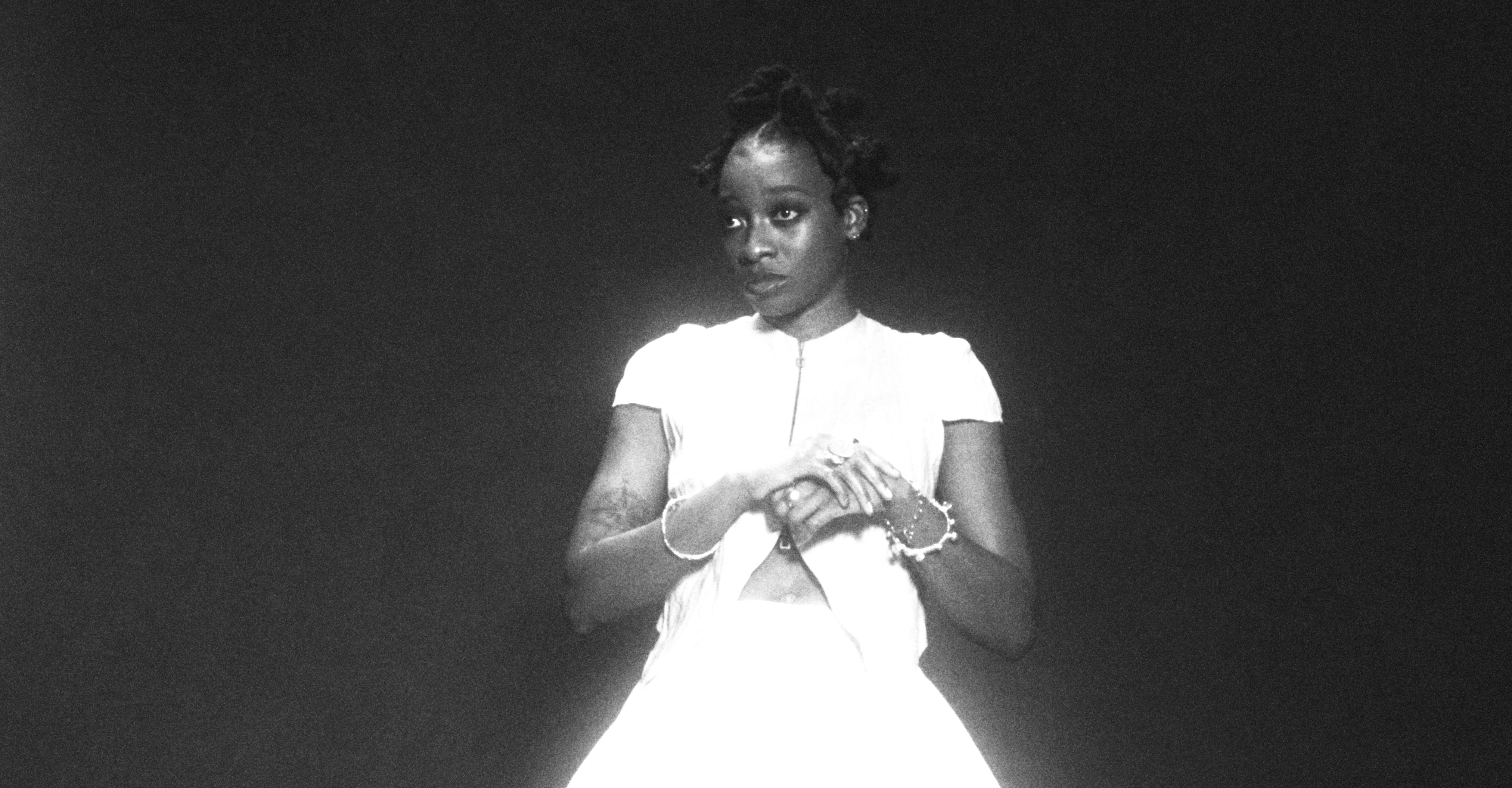 Little Simz in a white outfit stands against a dark background, looking to the side. The image is in black and white.
