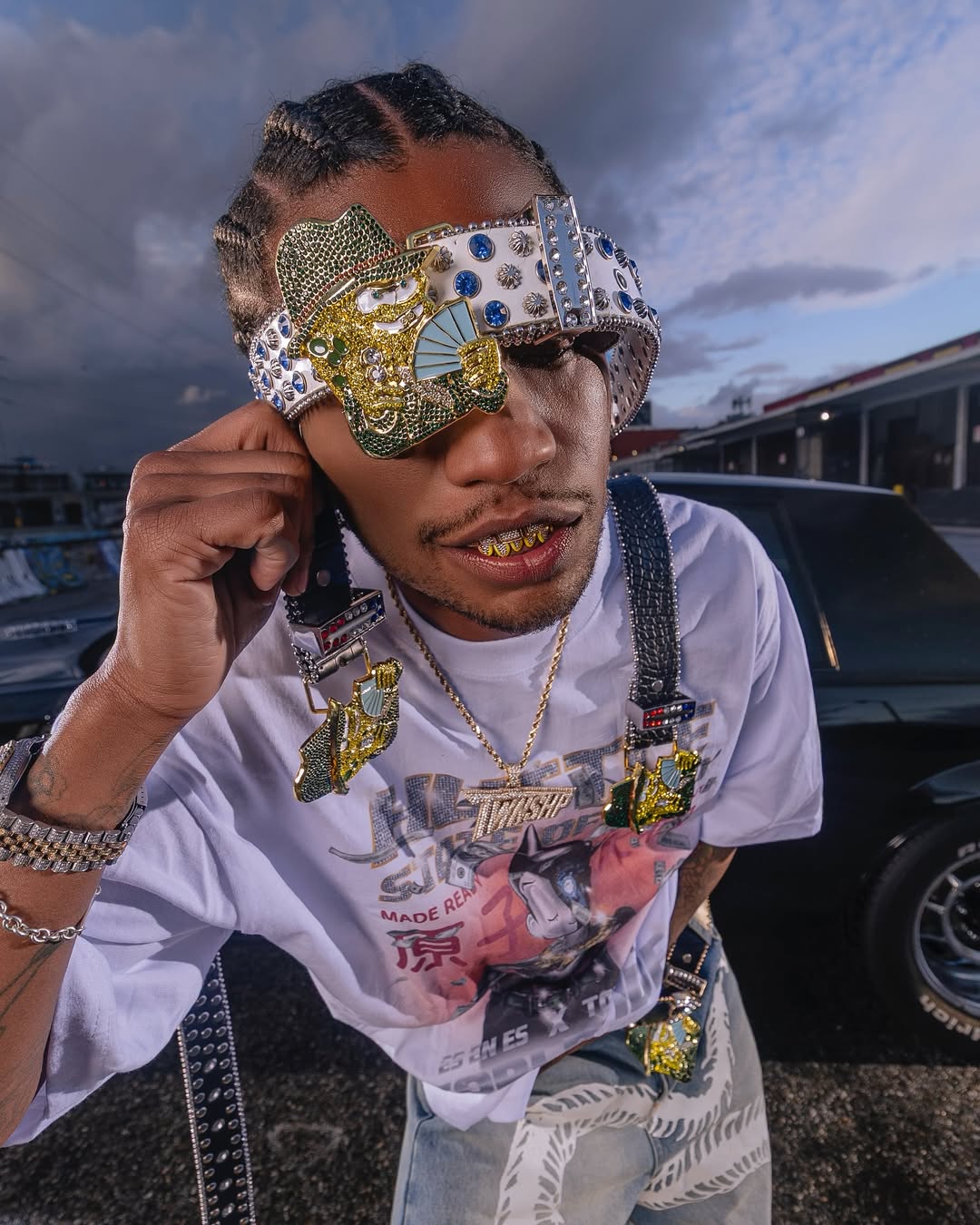 A person wearing a jeweled visor, gold grills, and a graphic t-shirt. They have braided hair and are standing near a car.