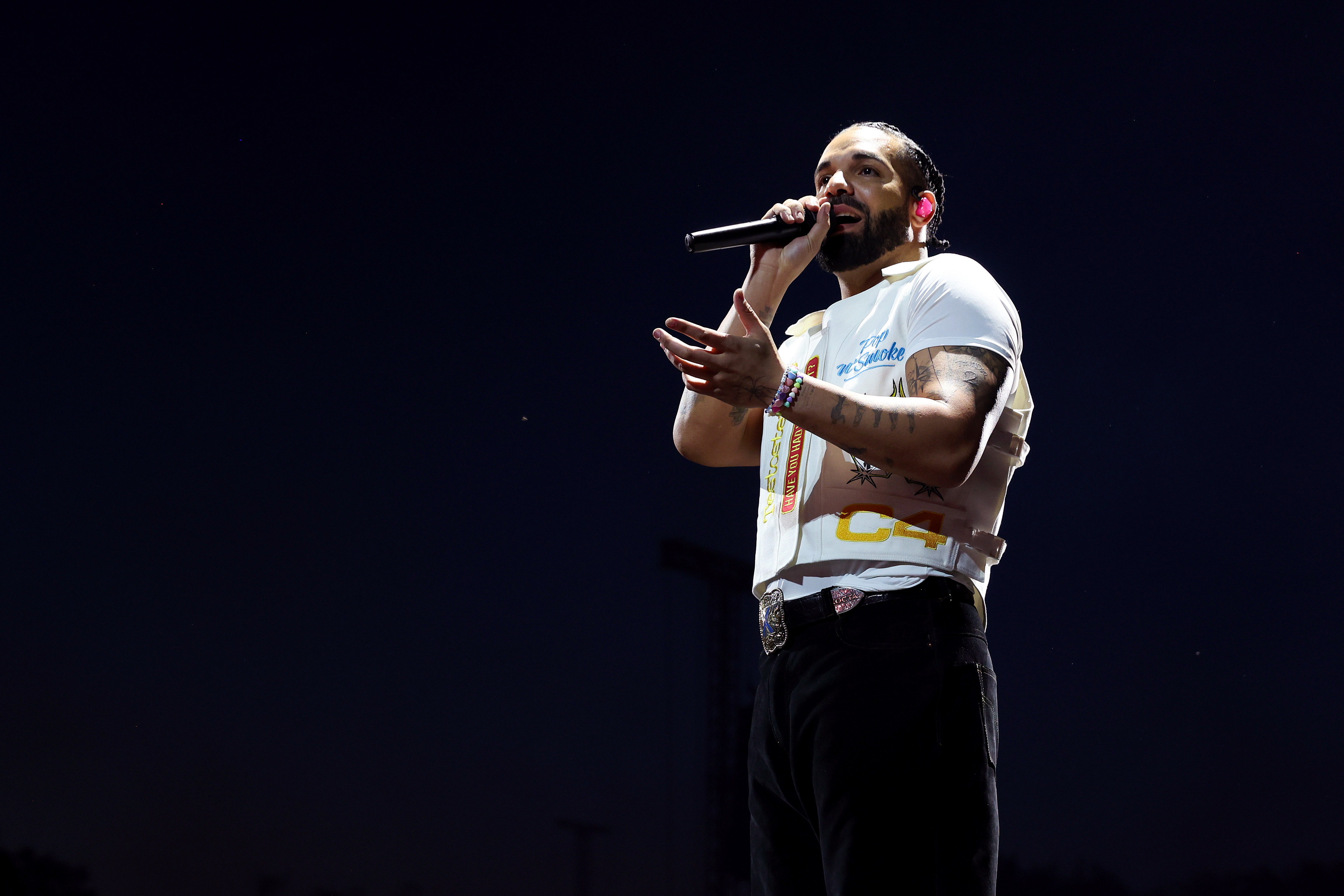 A man with a beard and braided hair performs on stage, holding a microphone, wearing a white t-shirt and black pants, against a dark background.