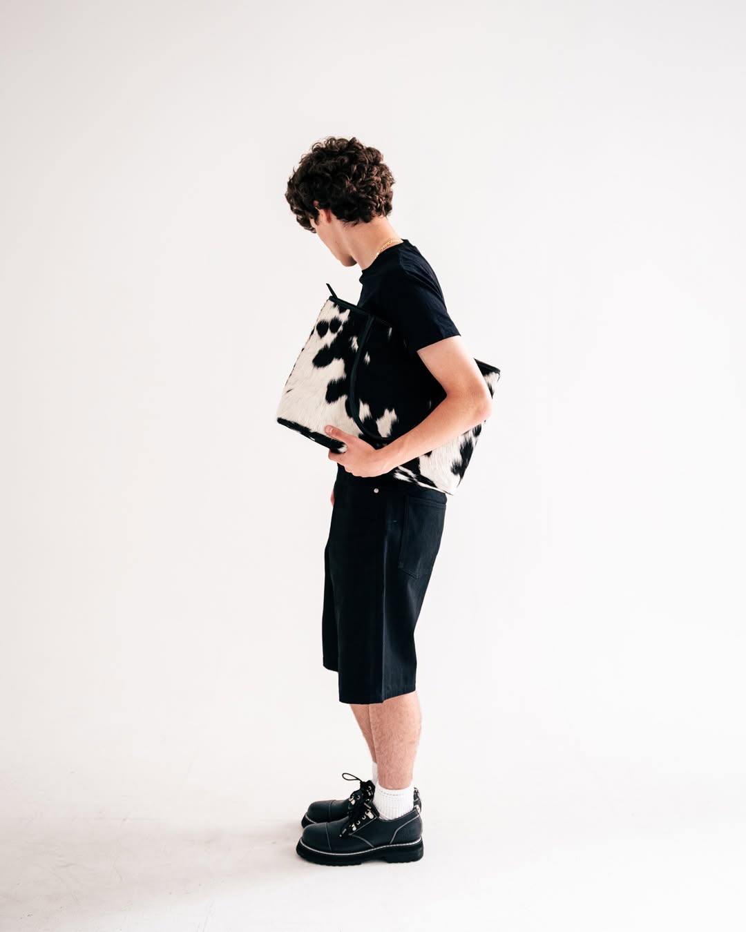 A person with curly hair, wearing a black outfit, holds a black and white patterned bag, standing against a plain background.