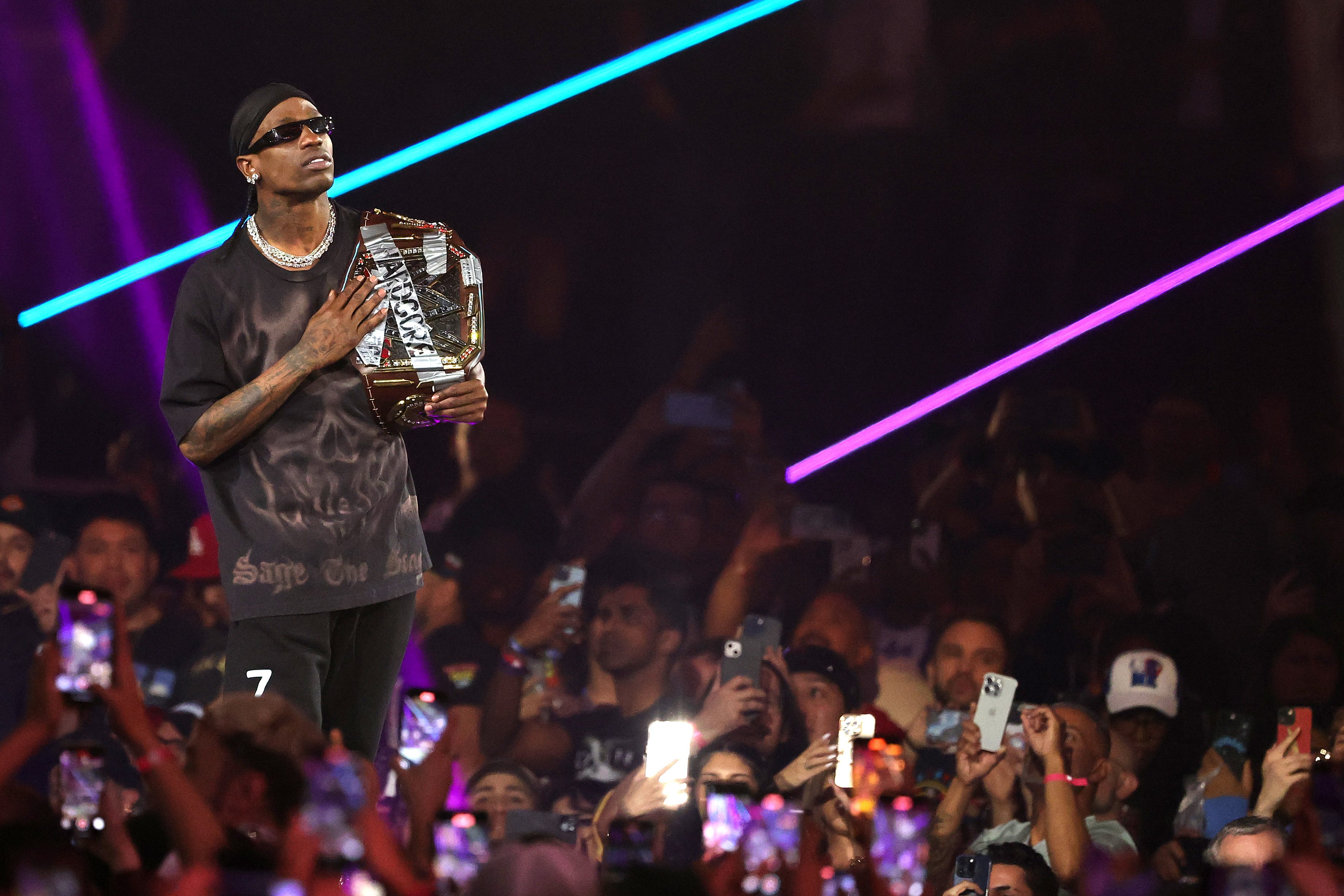 Travis Scott wearing sunglasses and a dark outfit holds a championship belt on stage, surrounded by a crowd with phones and colorful lights.