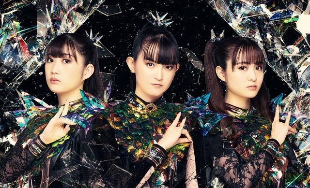 Three members of Babymetal in colorful, futuristic outfits with a dark, abstract background.