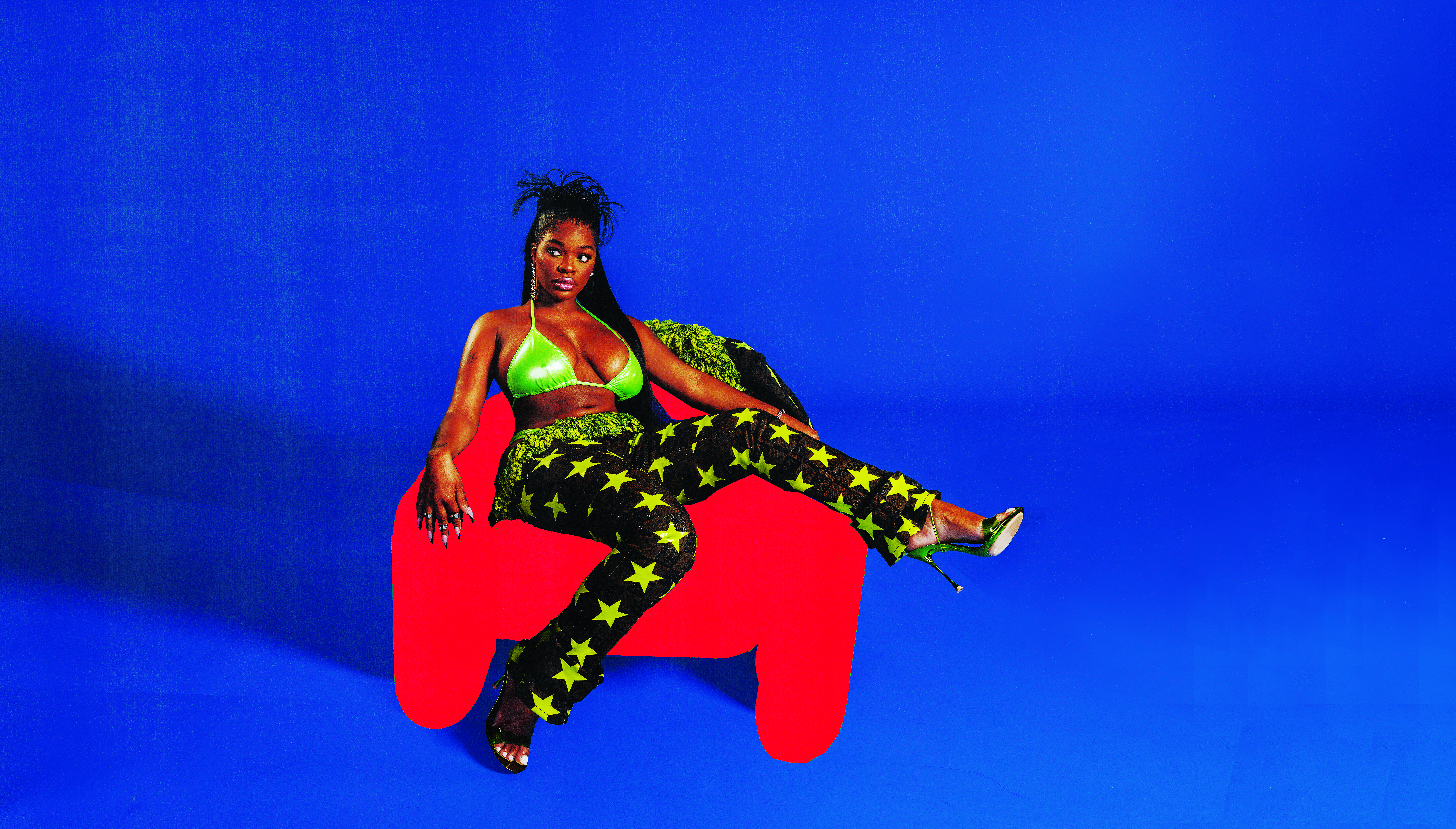 JT in a green top and star-patterned pants sits on a red chair against a blue background.