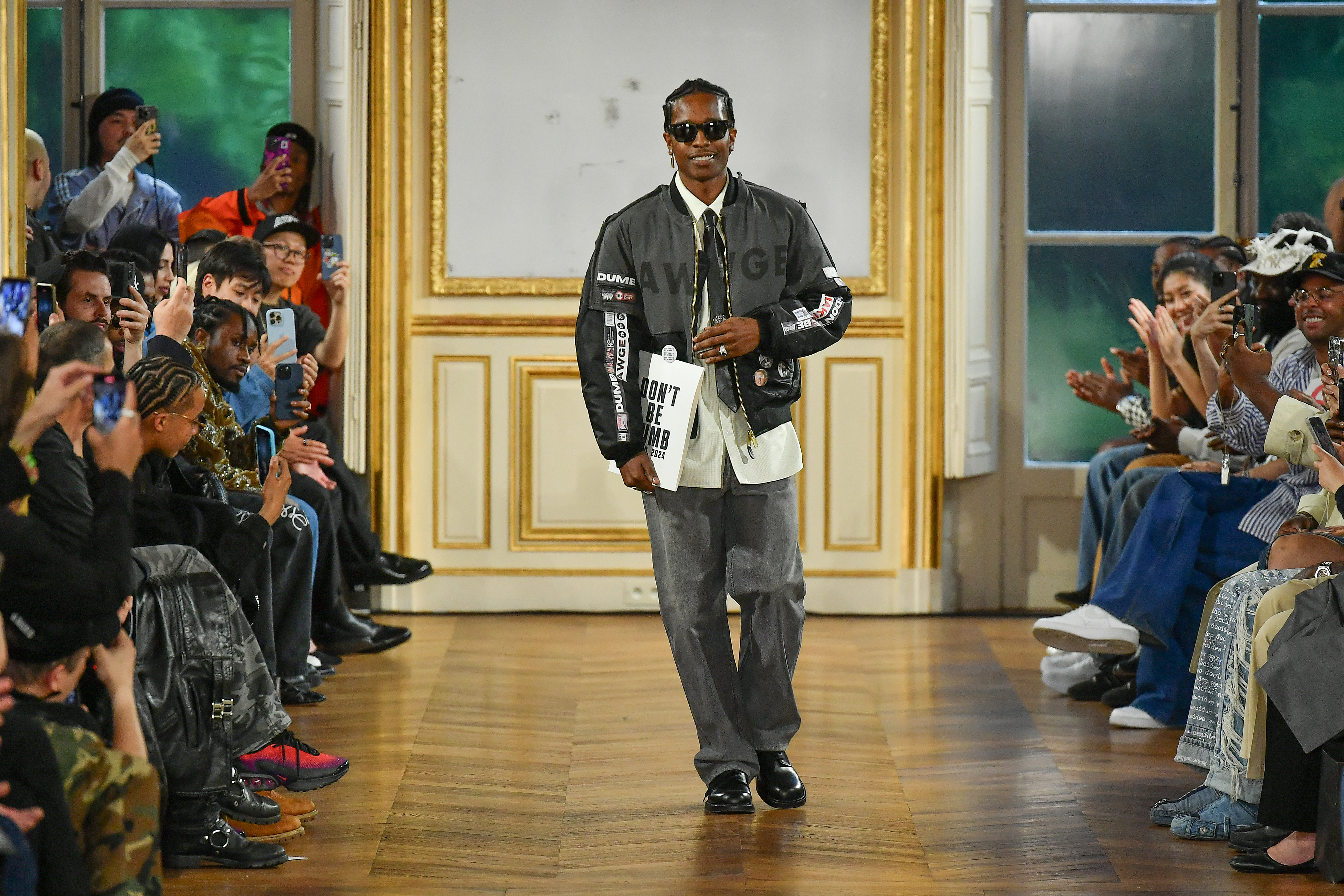 A$AP Rocky walks a fashion runway in a stylish gray outfit and sunglasses, holding a bag. The audience captures the moment with phones.