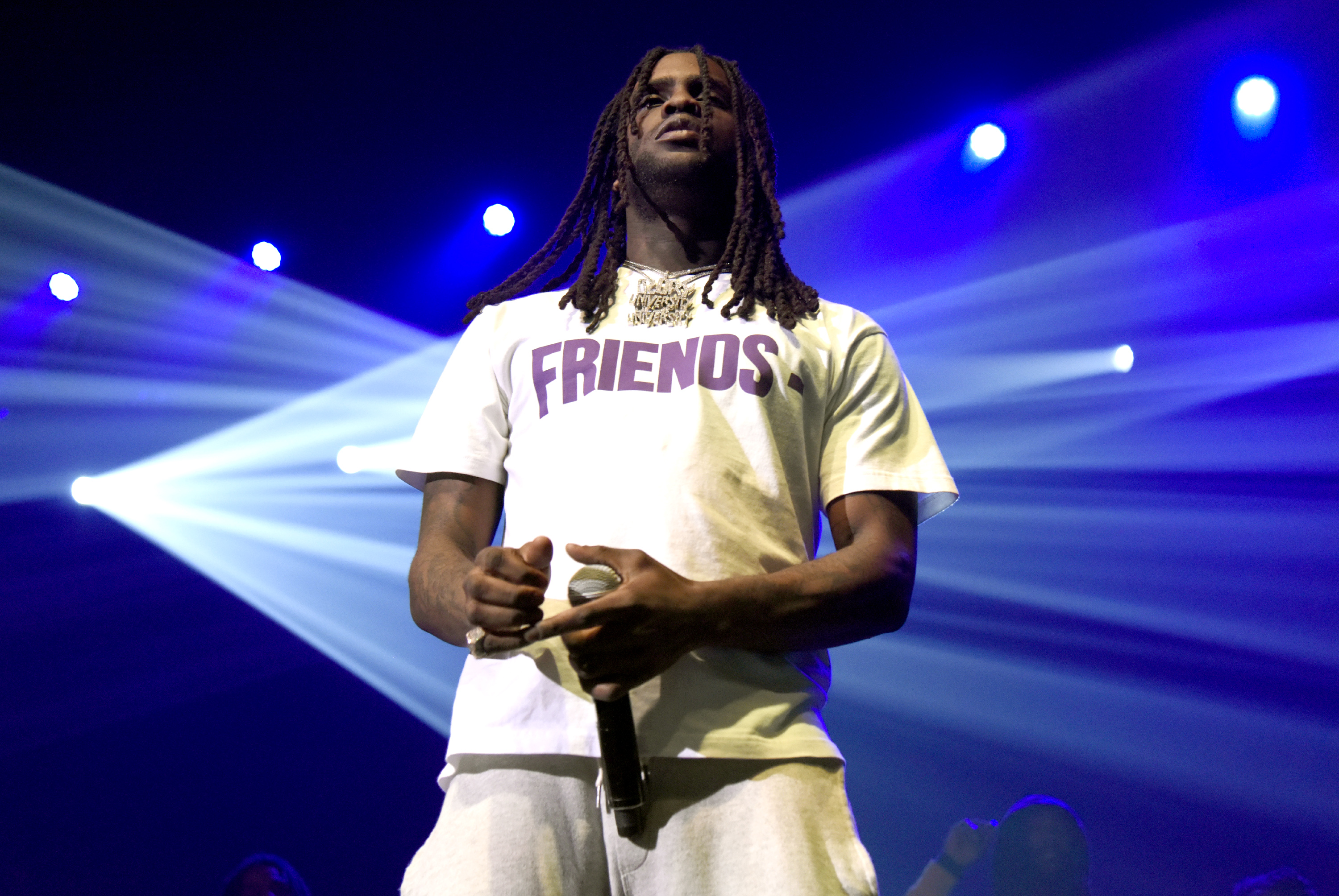 Chief Keef wearing a "FRIENDS" shirt, holding a microphone, with dramatic blue and white stage lighting.