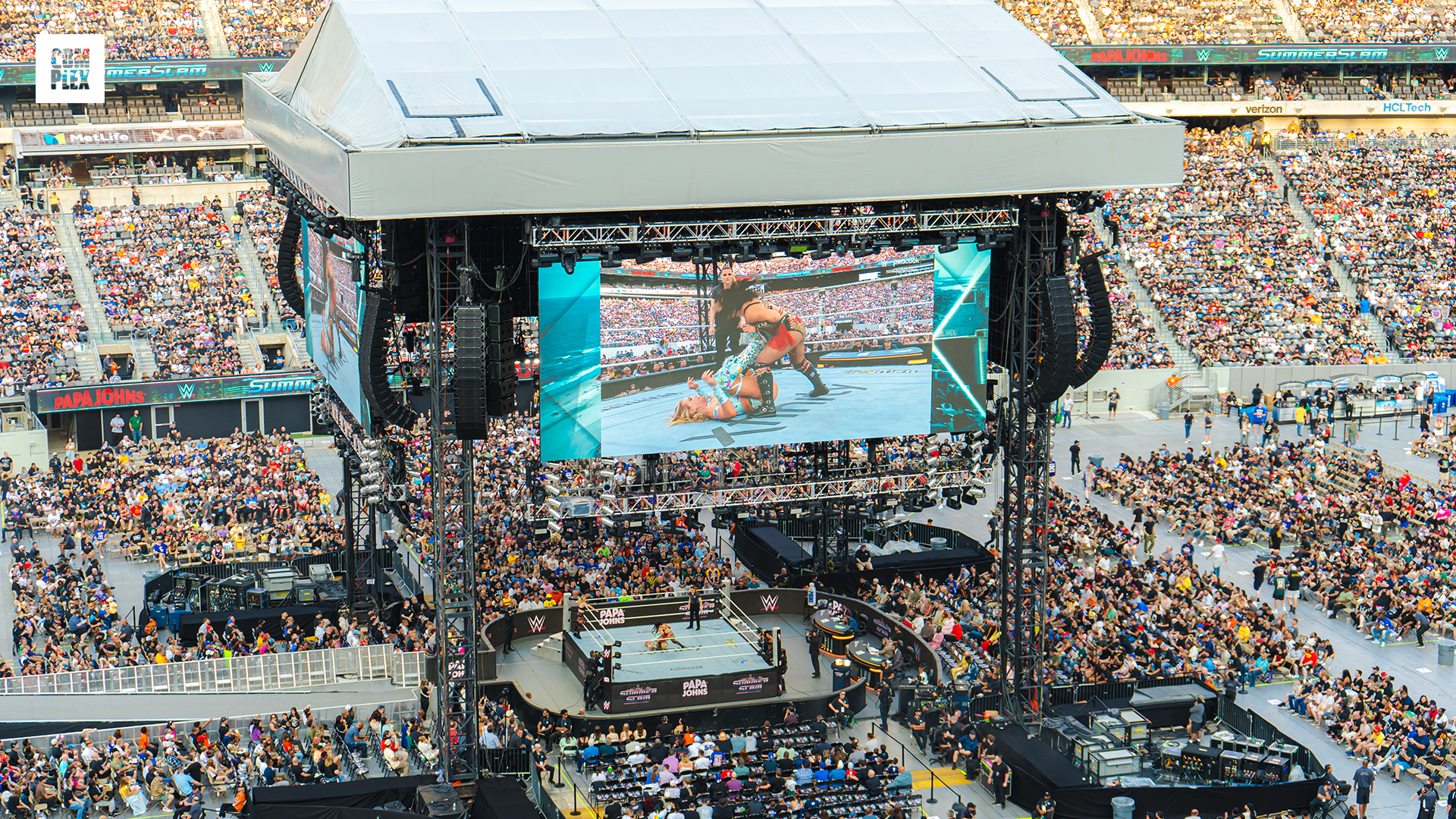 A large wrestling event in a stadium, with a crowded audience and a big screen displaying the match.