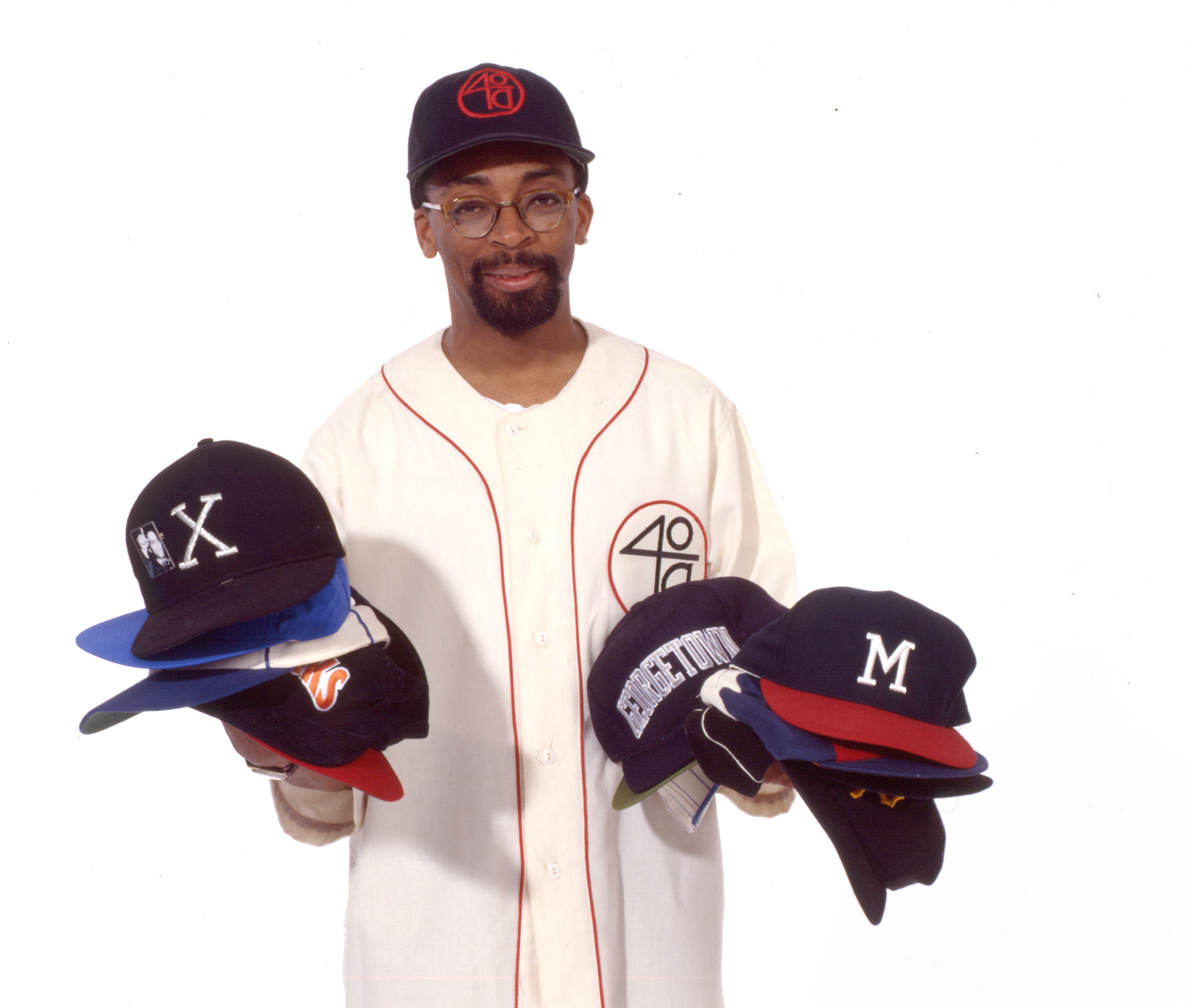 A man wearing a baseball jersey and cap, holding multiple caps in each hand.