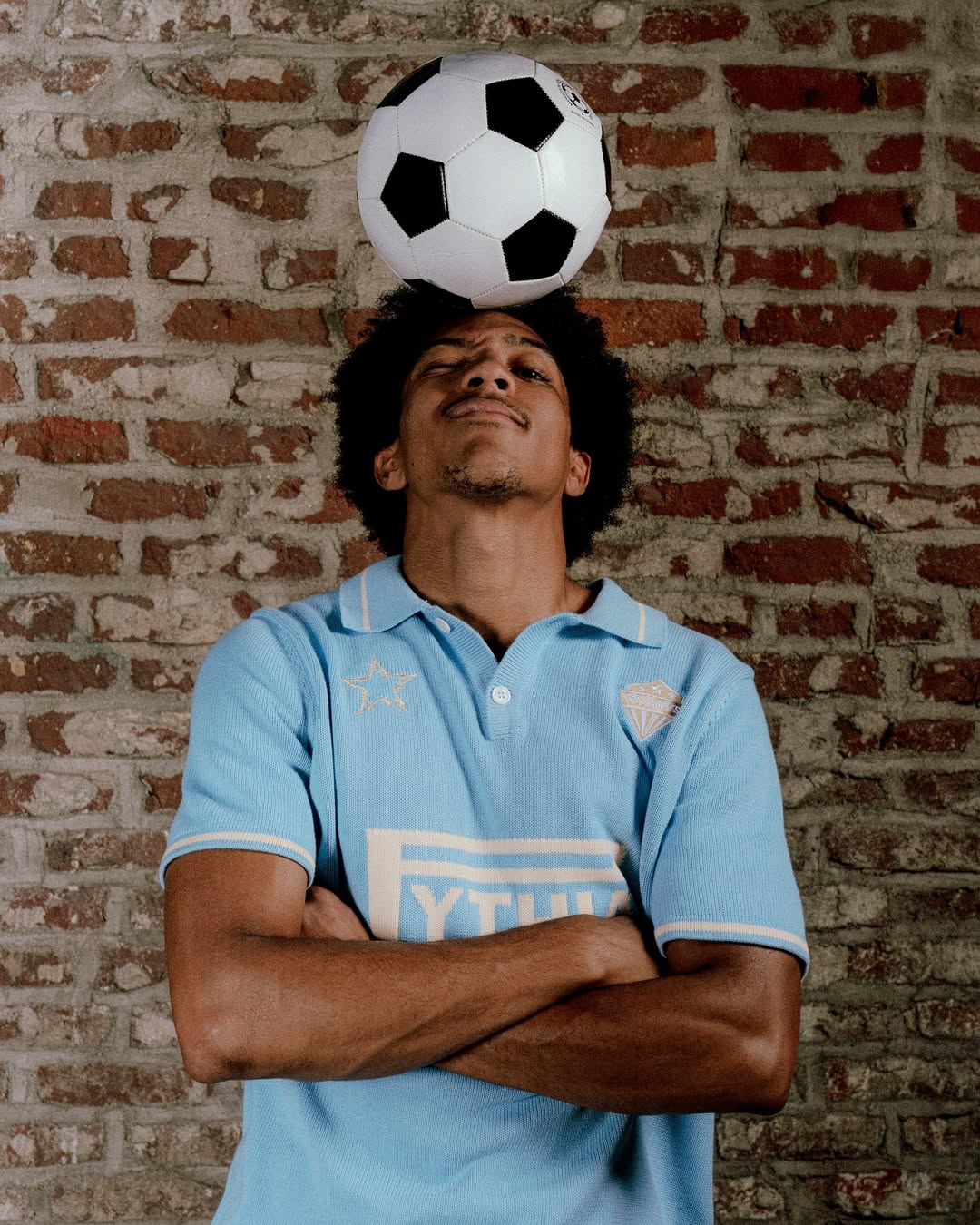 A person in a blue shirt balances a soccer ball on their head against a brick wall, arms crossed and eyes closed.