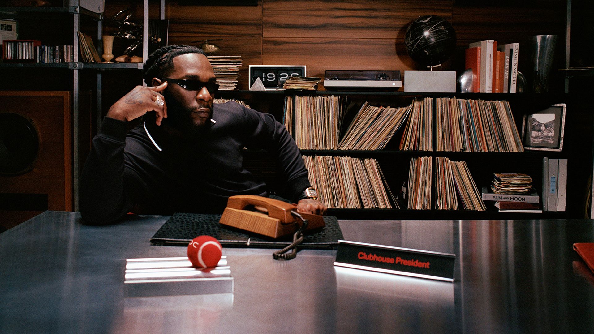A man in sunglasses sits at a desk labeled "Clubhouse President," surrounded by shelves of records and books, with a retro phone.