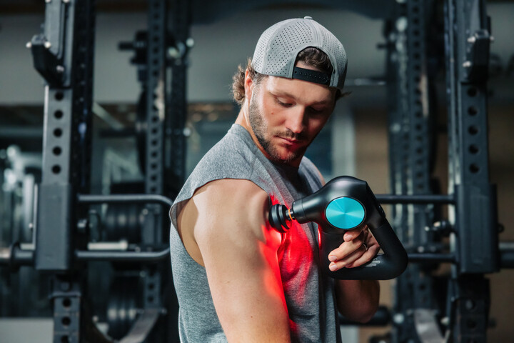 Man using a massage gun on his arm in a gym, wearing a gray sleeveless shirt and cap. Gym equipment is visible in the background.