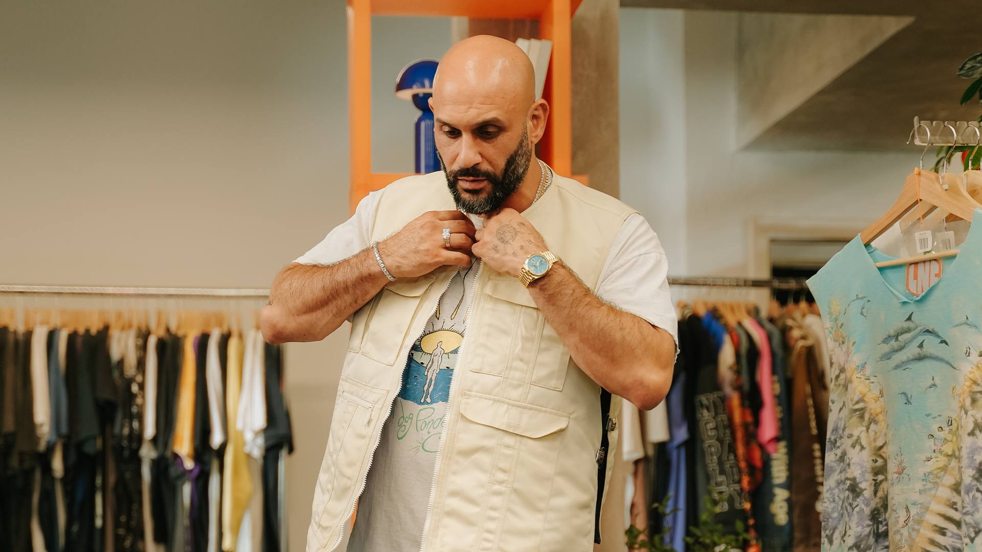 A man with a beard and bald head adjusts a beige vest in a clothing store. Various clothes are displayed on racks around him.