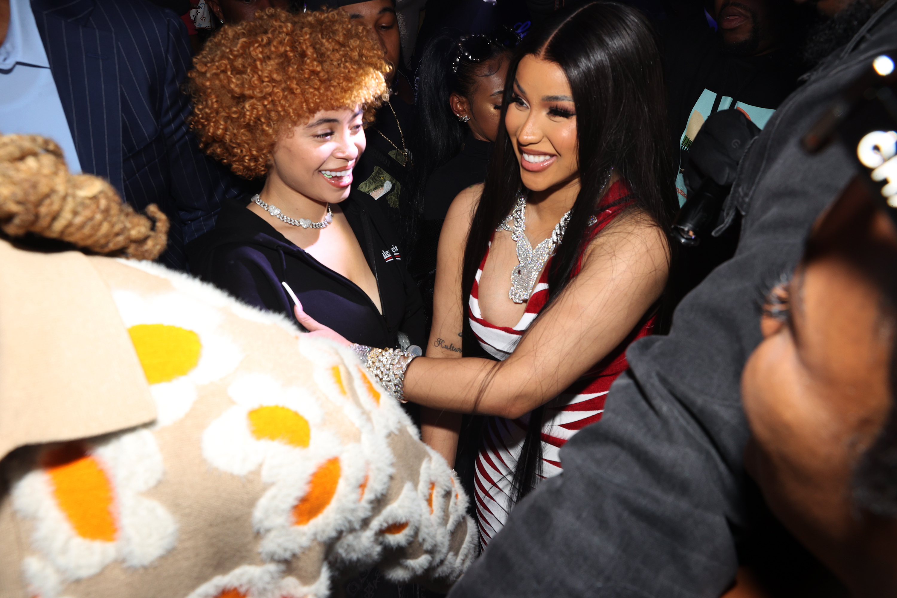 Ice Spice and Cardi B smiling and interacting at an event, surrounded by people. Ice Spice has curly hair, and Cardi B wears a red-striped outfit.