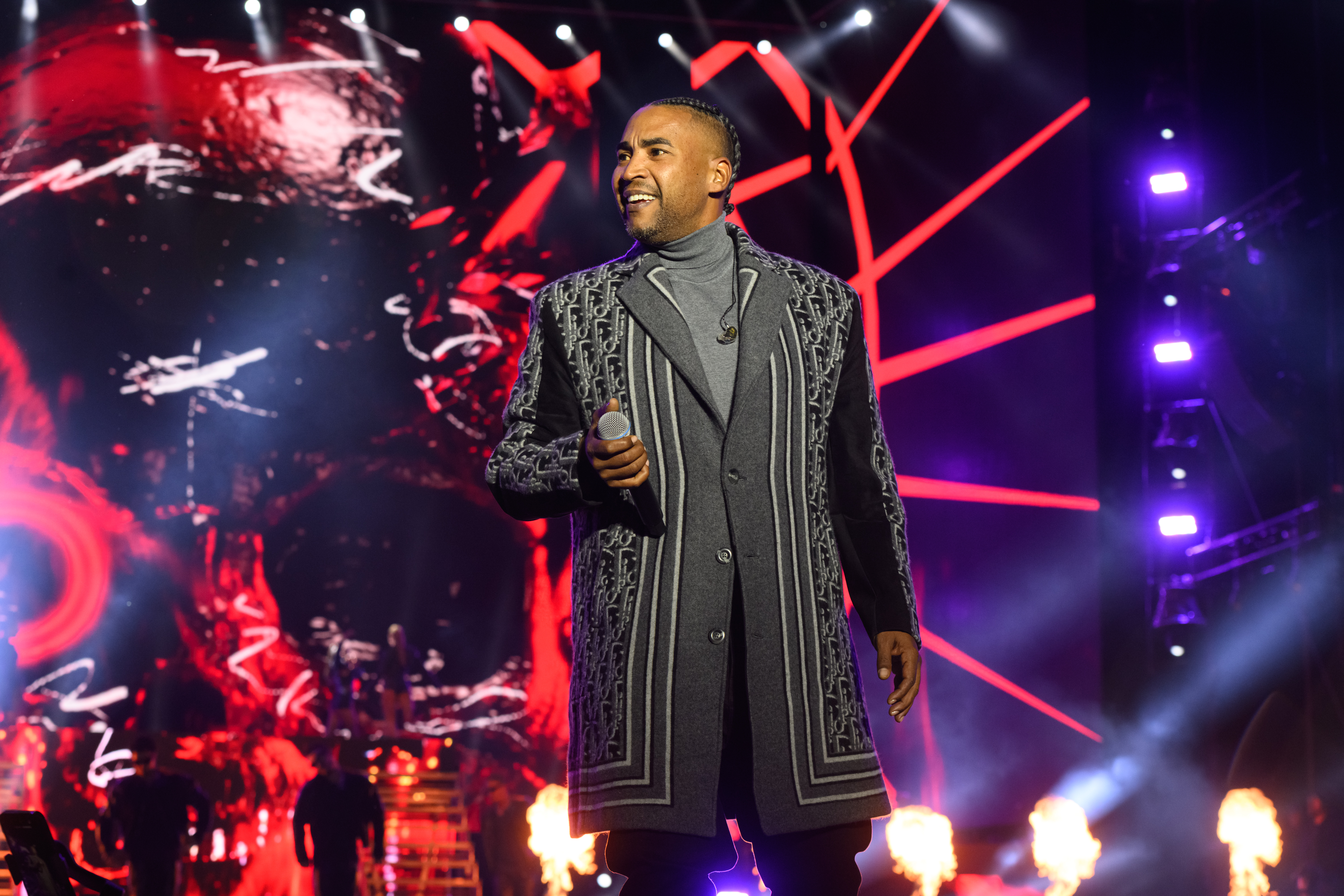 Don Omar on stage holding a microphone, wearing a patterned coat, with a vibrant red and black background and stage lights.