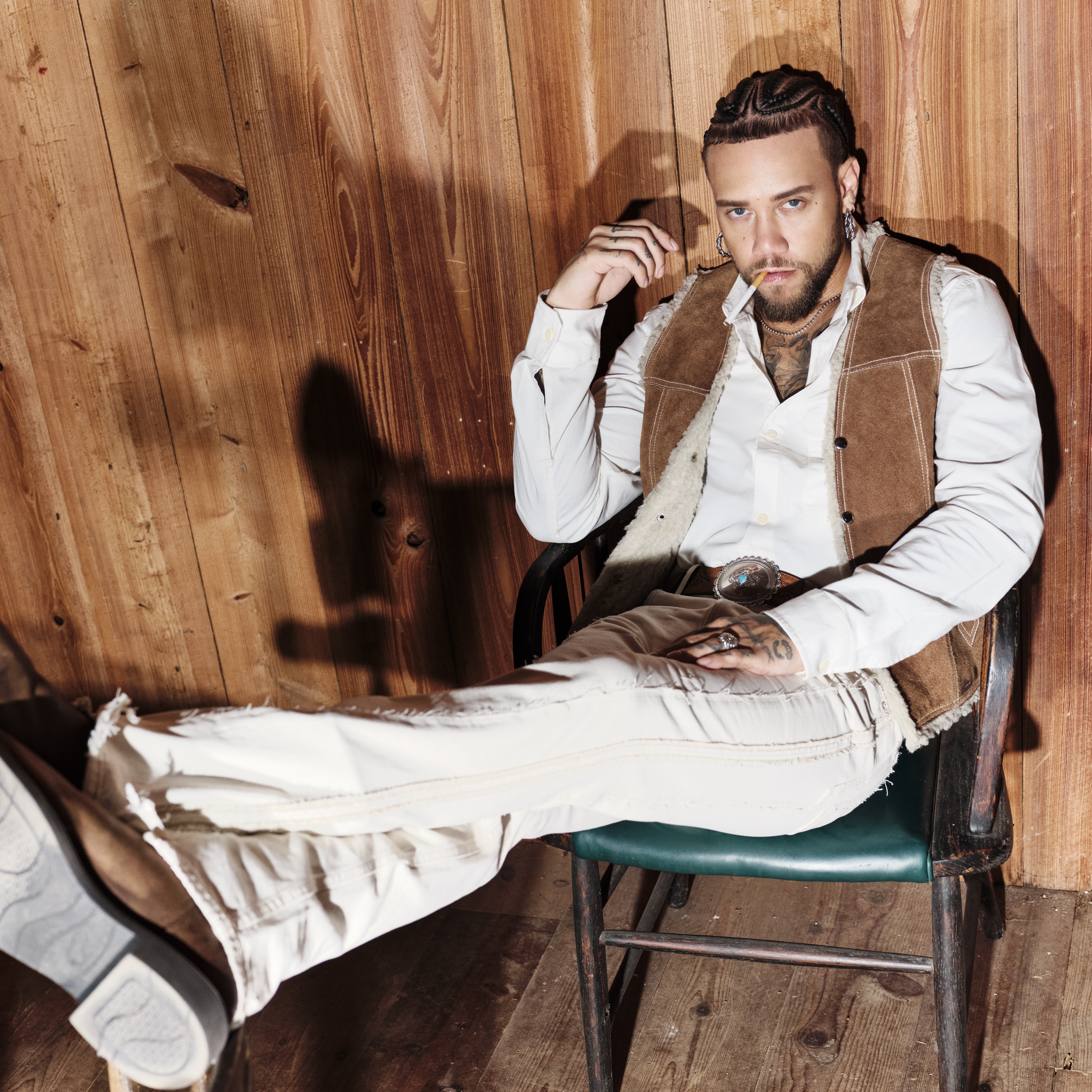 A man with braided hair, wearing a brown vest and white shirt, sits in a chair against a wooden wall, legs stretched out.
