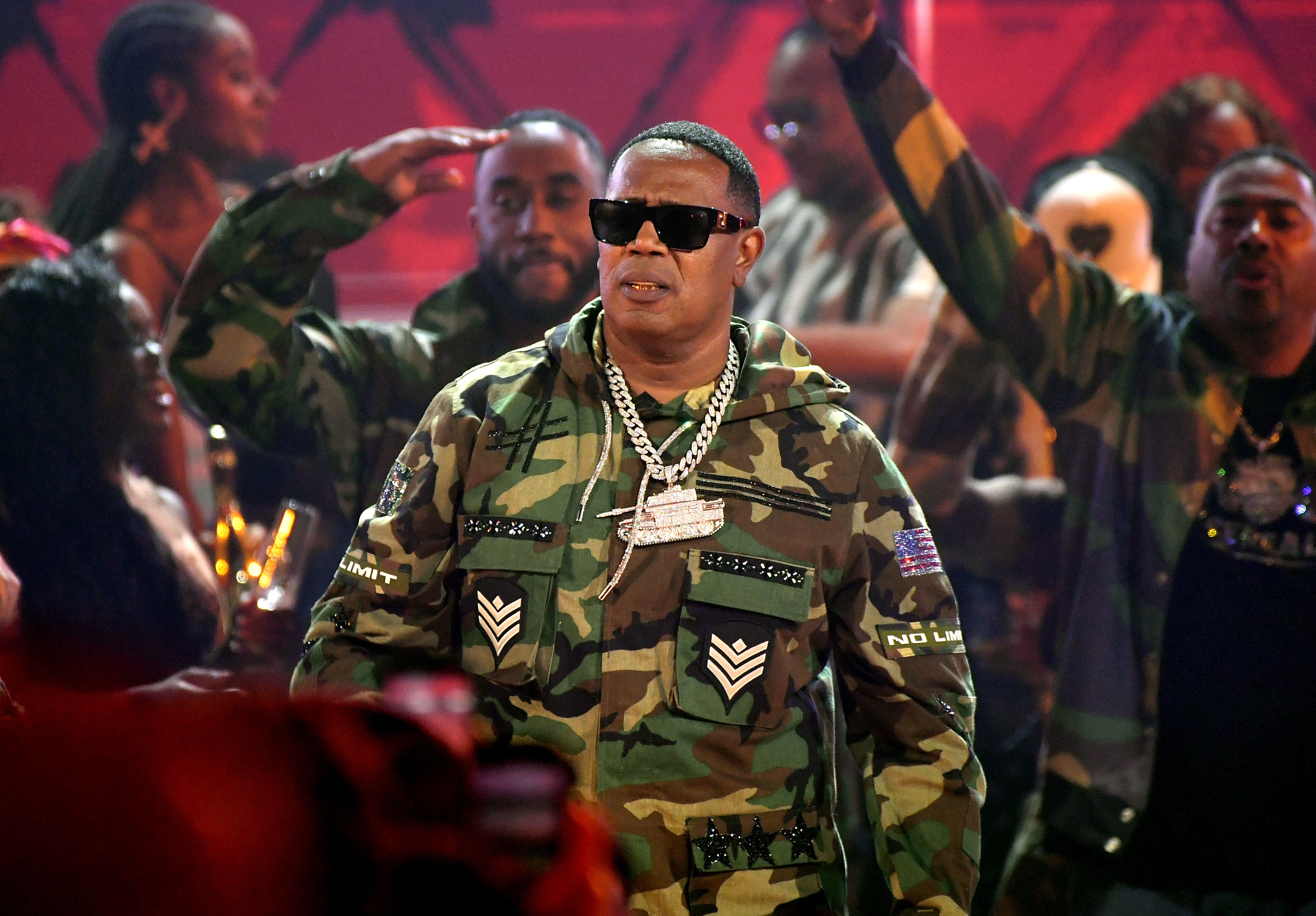 Master P in camo outfit, wearing sunglasses and a chain, surrounded by people in similar attire on stage.