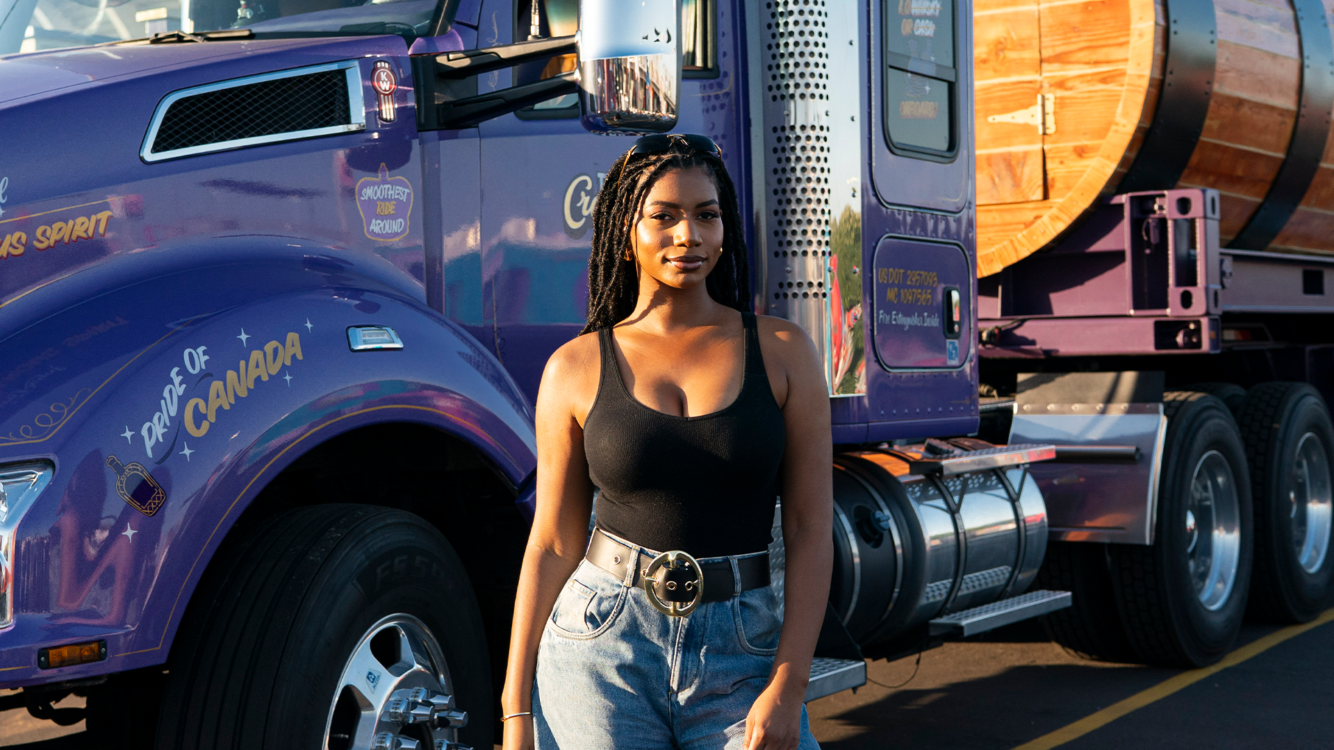 A woman stands in front of a purple truck with "Pride of Canada" written on it. She wears a black tank top and jeans.