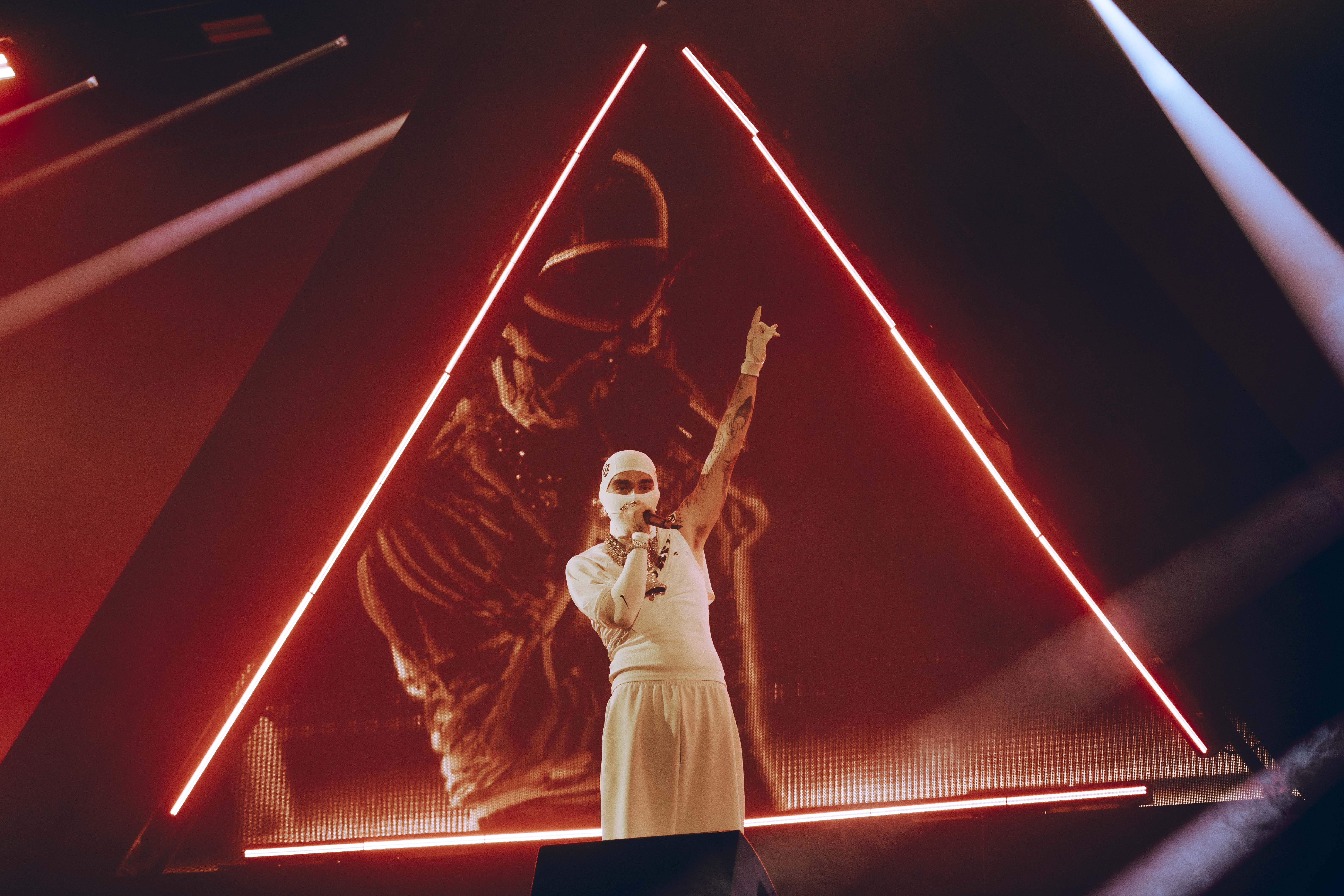 A performer wearing a white outfit and mask sings on stage, with a triangular red light structure in the background.