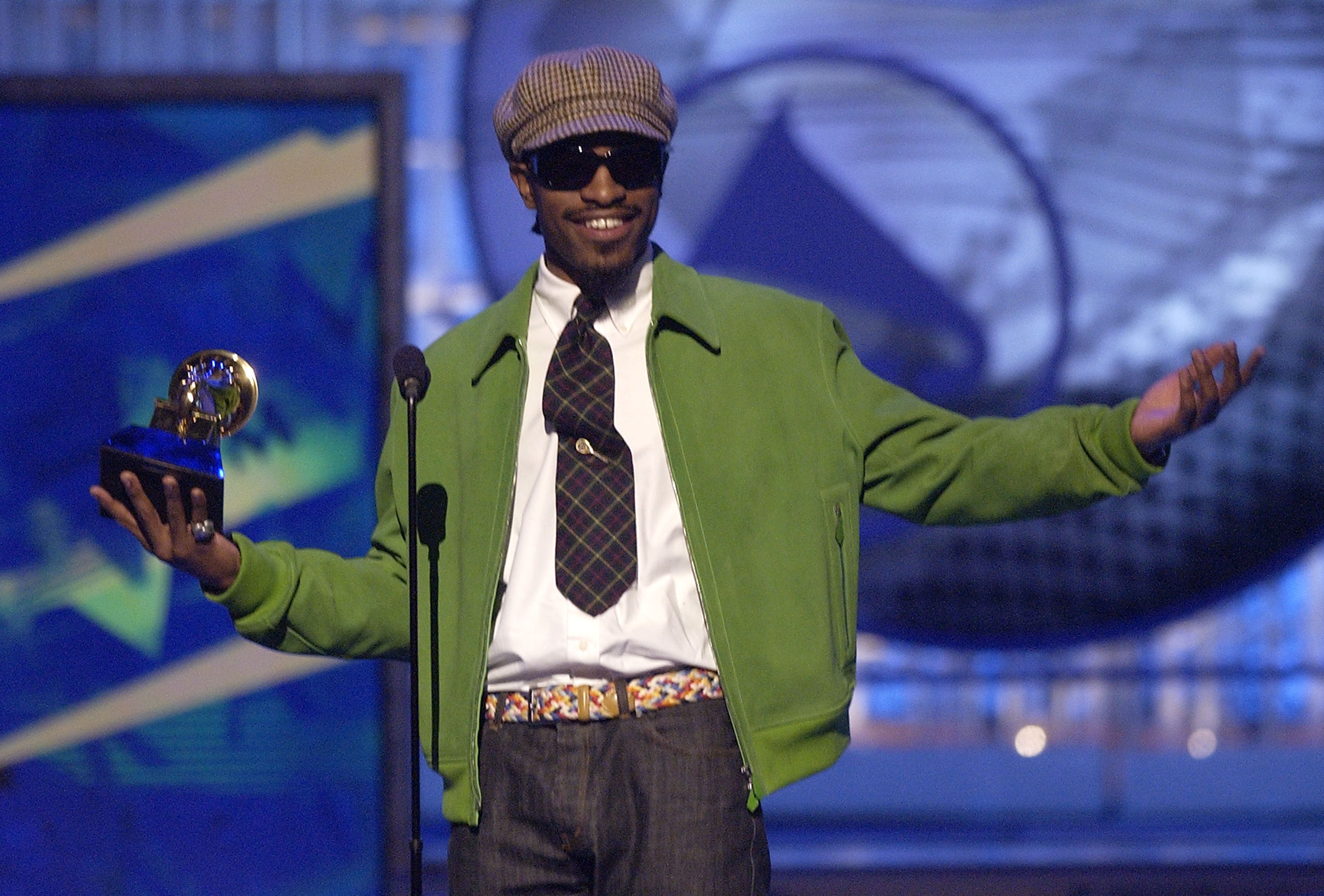 Andre 3000 in a green jacket and hat, holding a Grammy award, speaking at a podium.