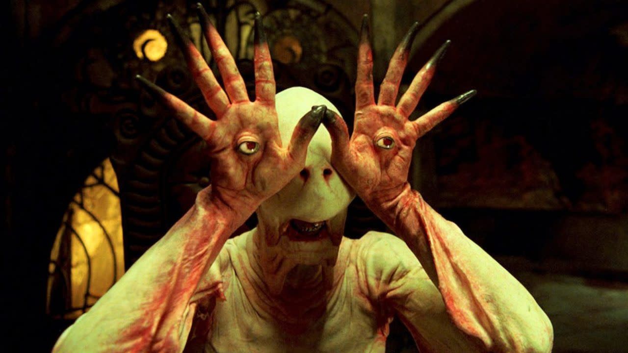 A creature with pale skin and eyes on its hands, from "Pan's Labyrinth," stands in a dimly lit room.