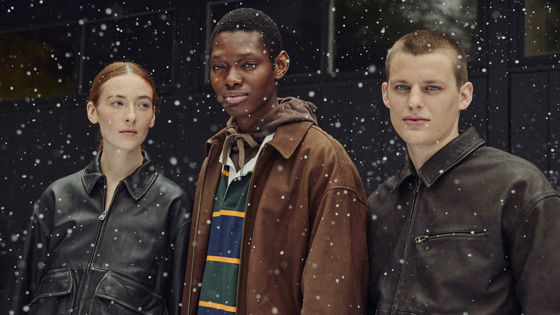 Three models in winter clothing stand outside in falling snow, wearing jackets and looking at the camera.