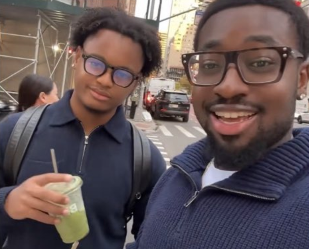 Two people with glasses smiling on a city street. One holds a green drink. Background shows buildings and a street corner.
