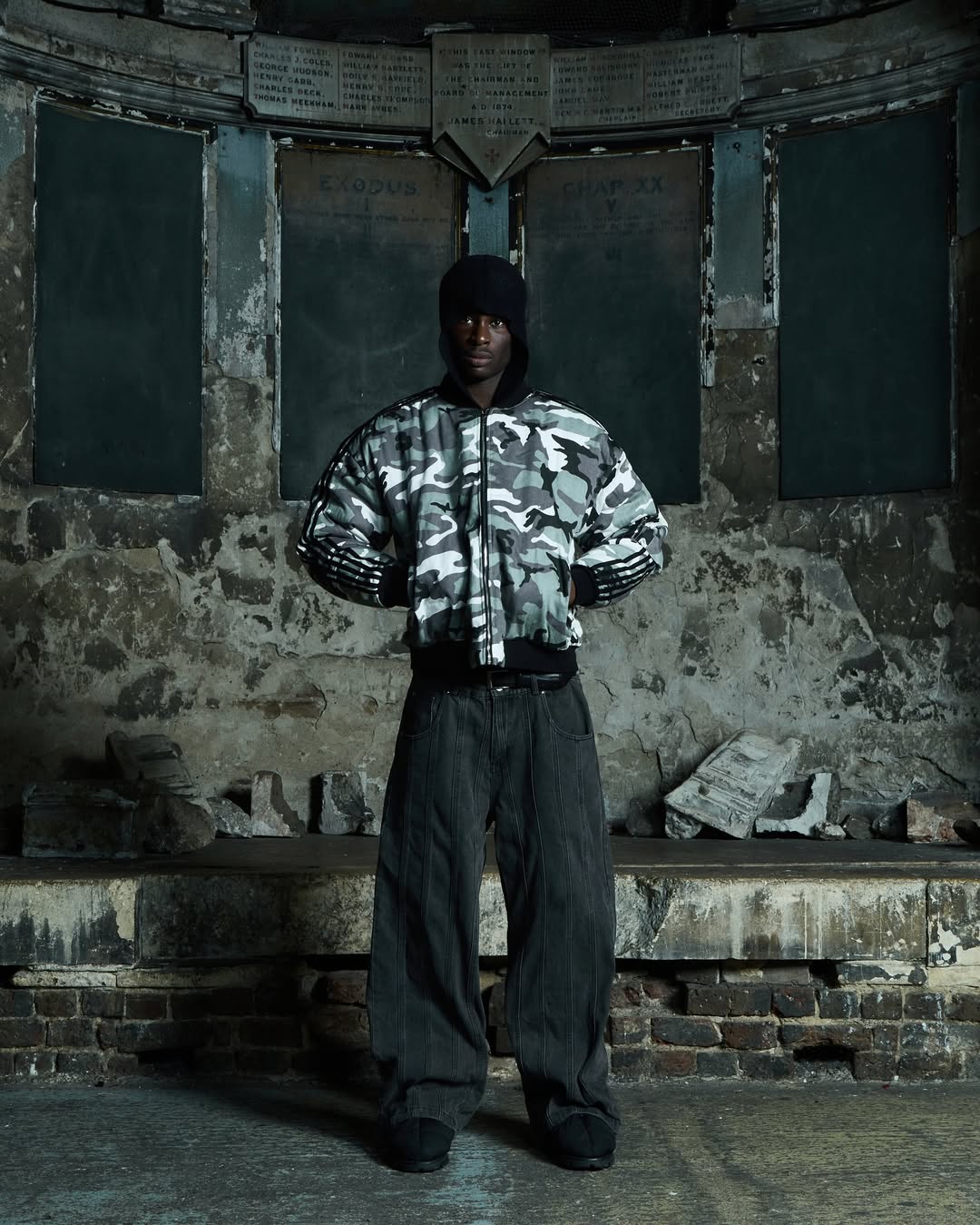 A person in a camouflage jacket and dark pants stands in a worn, stone interior with a historical or rustic backdrop.