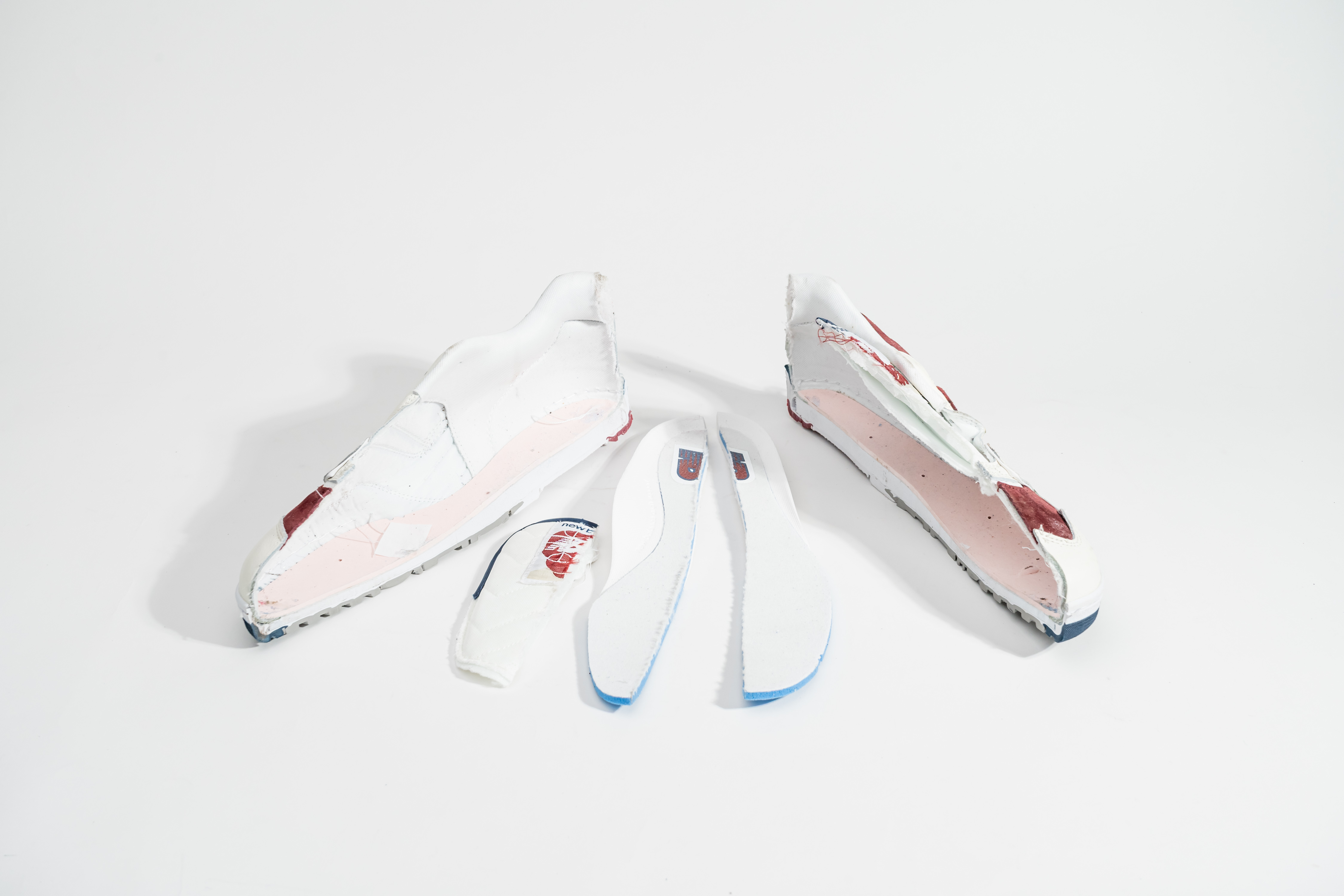 A pair of deconstructed white sneakers with visible layers, including pink and blue components, displayed on a plain background.