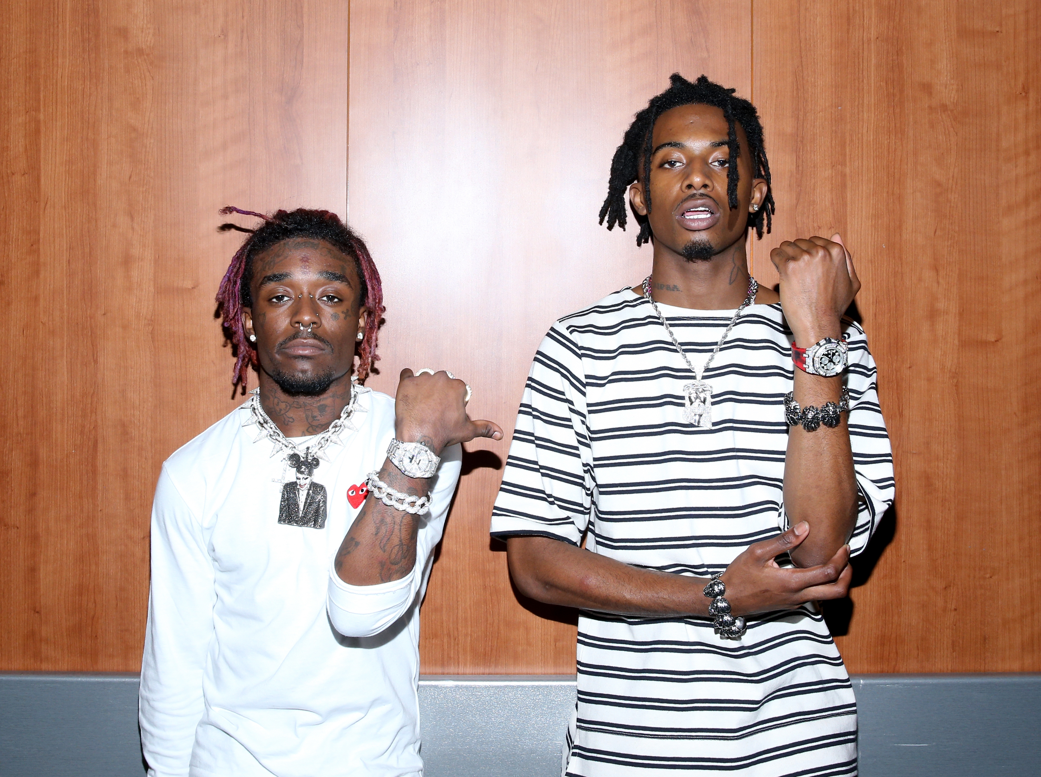 Lil Uzi Vert and Playboi Carti pose together, showcasing jewelry, against a wooden backdrop.