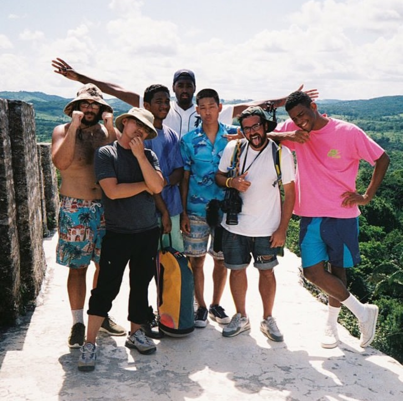 A group of seven men posing on a sunny day with a scenic landscape in the background. One holds a camera, and others wear casual summer attire.